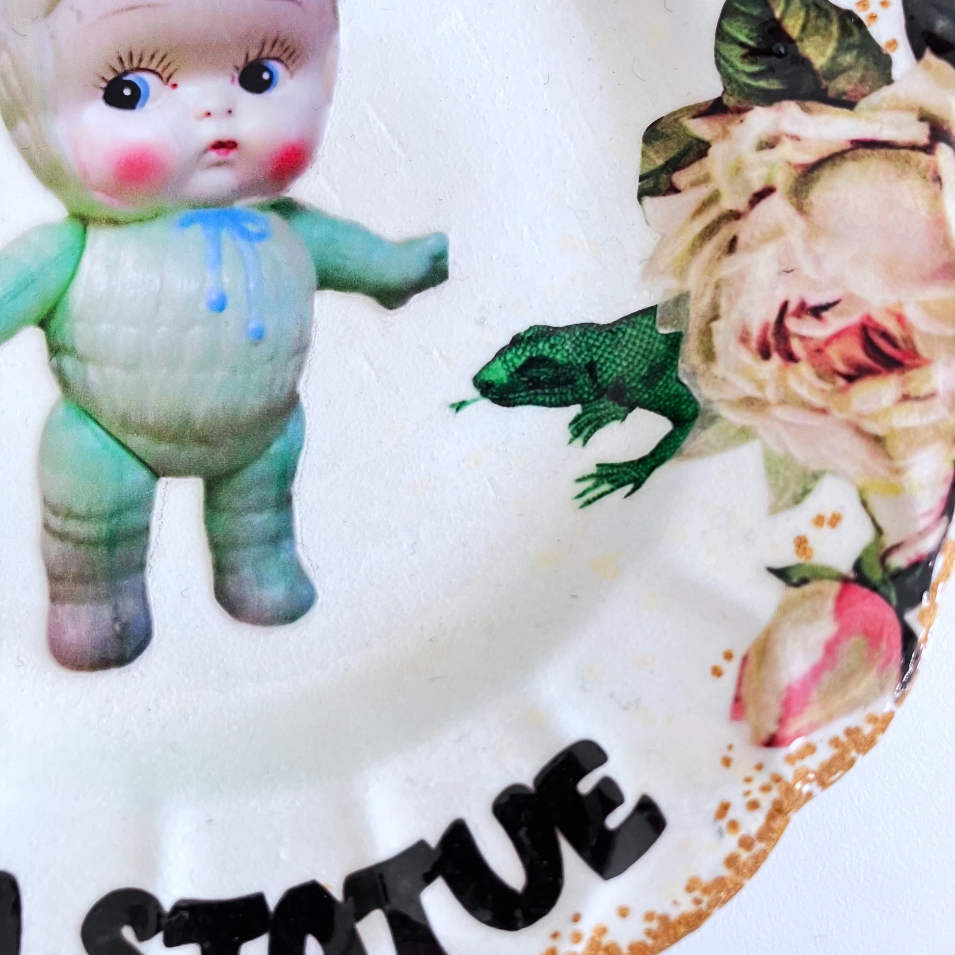 "As Unmoved As A Statue" white wall plate by House of Frisson, featuring a kitsch vintage plastic doll with roses and House of Frisson's fly and lizard. Closeup detail of the collage. 
