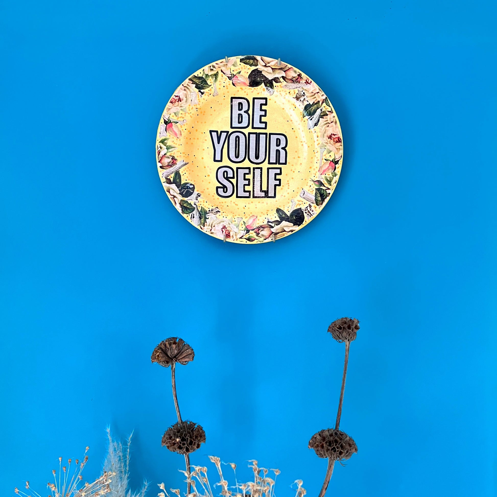 Yellow Upcycled Wall Plate by House of Frisson "Be Yourself". Featuring a collage with vintage flowers and bones. Wall plate hanging on a blue wall.