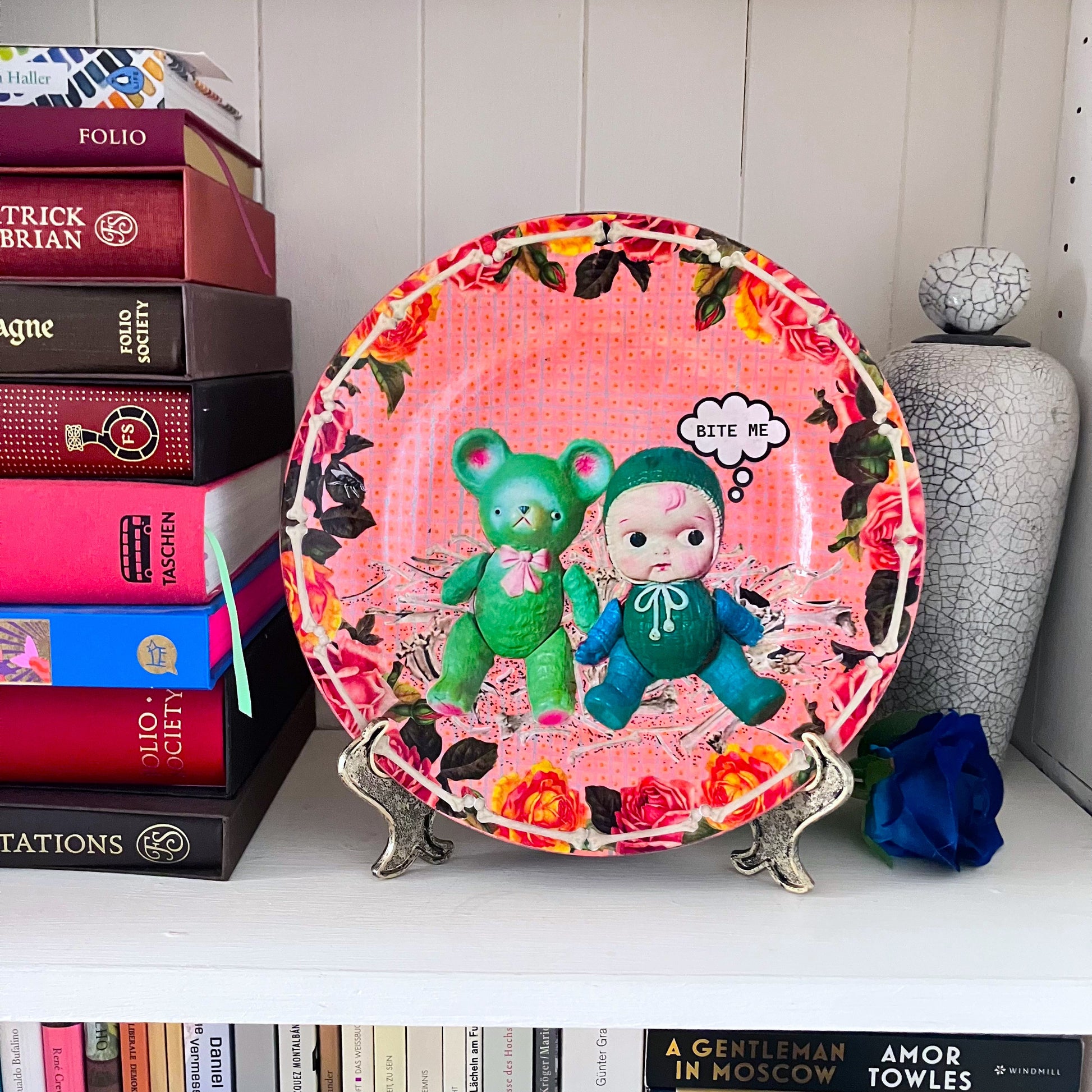 Peach Pink upcycled wall plate from House of Frisson, featuring a vintage celluloid bear and a doll, surrounded by bones, in a frame of colourful roses and bones. Plate on a plate hanger resting on a shelf.