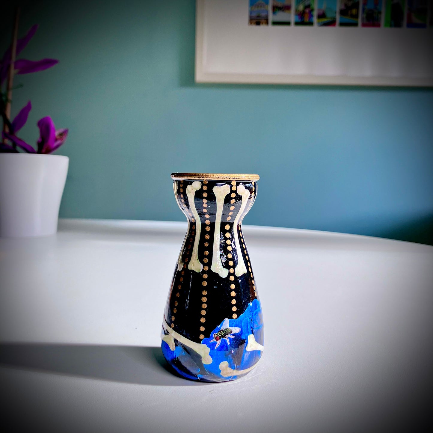 Black Upcycled Glass Bottle/ Vase by House of Frisson, featuring a collage of human bones and blue roses against a black background with some golden dots accent. Quirky and cool.