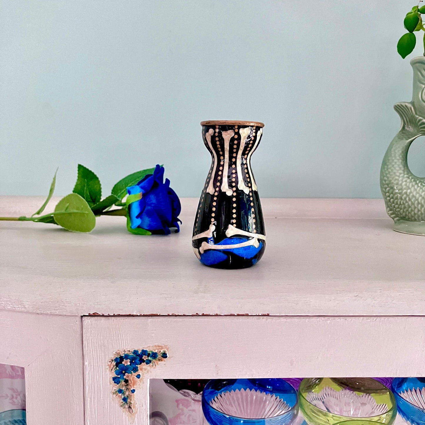 Black Upcycled Glass Bottle/ Vase by House of Frisson, featuring a collage of human bones and blue roses against a black background with some golden dots accent. Quirky and cool.