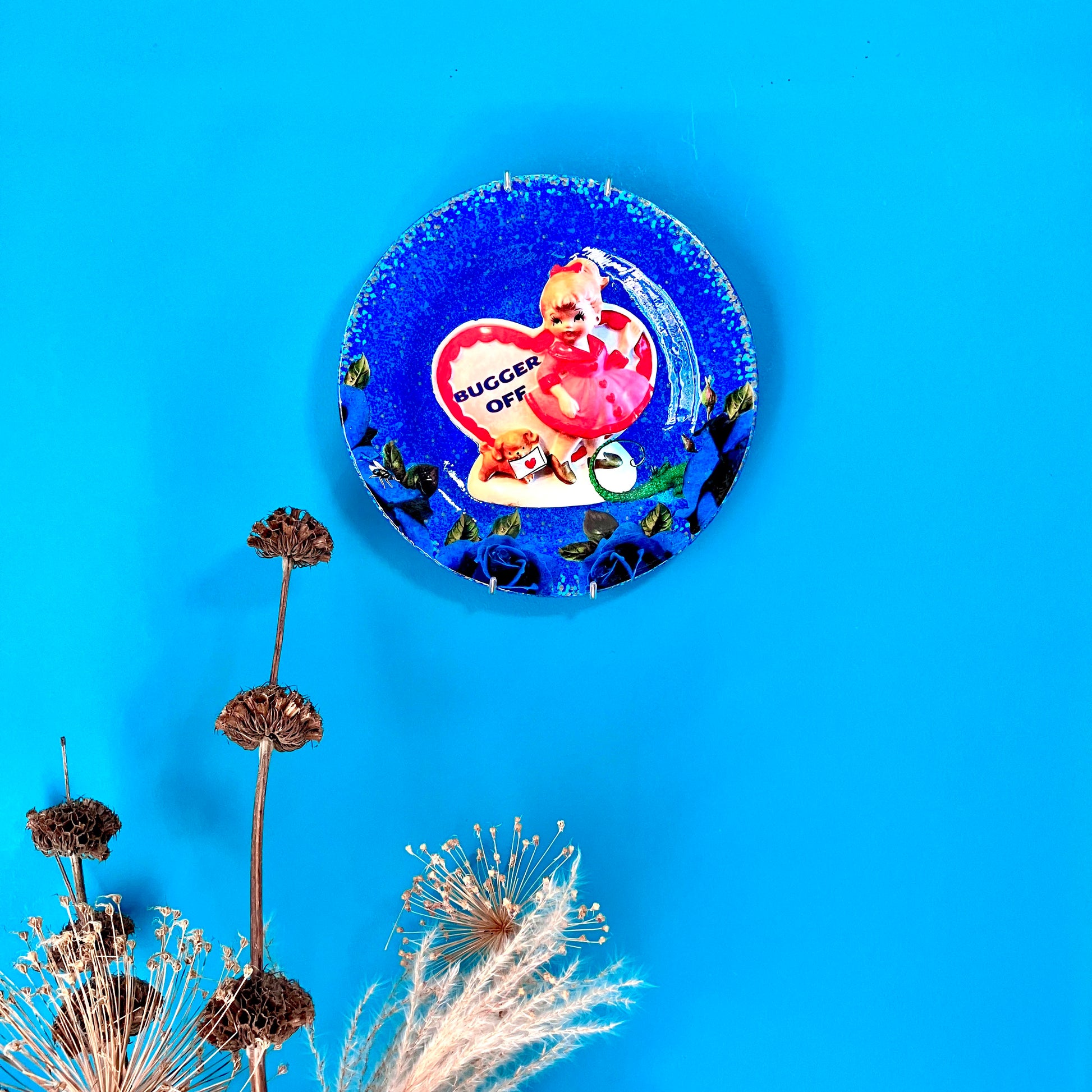 "Bugger Off" blue Wall Plate by House of Frisson, featuring a kitsch vintage porcelain figure among blue roses and a lizard and a fly. Plate hanging on a blue wall.