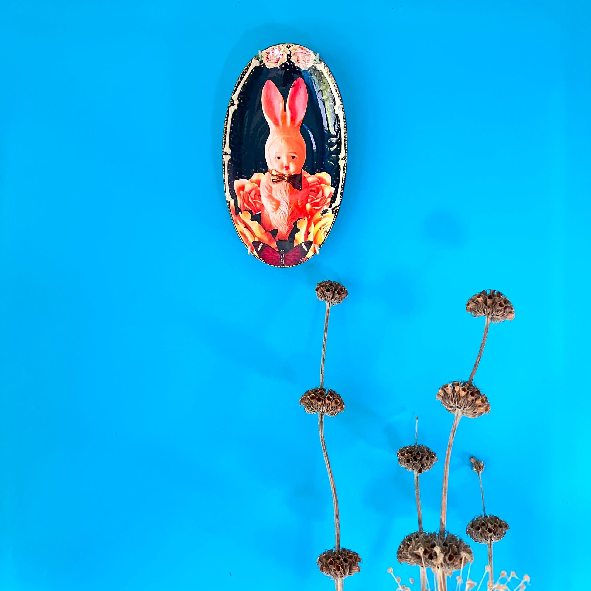 Black upcycled wall plate by House of Frisson, featuring a kitsch vintage squeaky bunny image surrounded by roses and bones. Plate hanging on a blue wall.