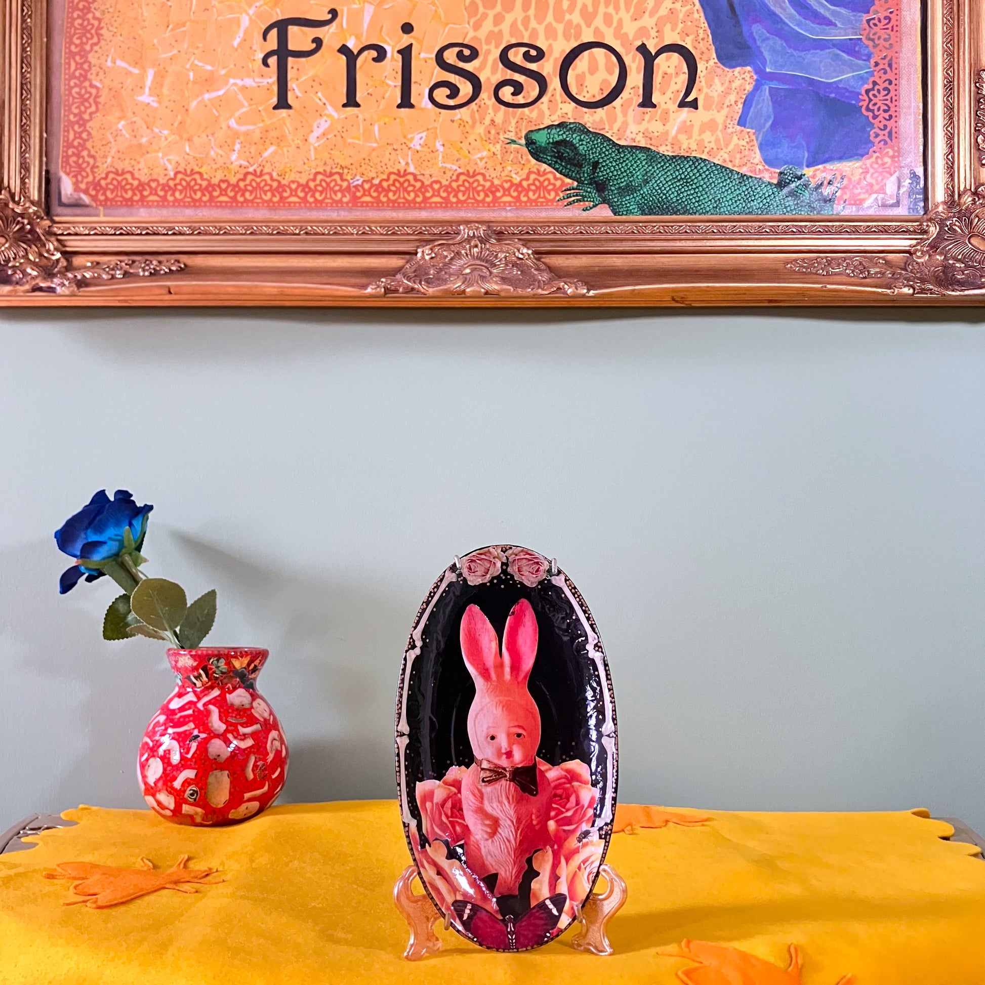 Black upcycled wall plate by House of Frisson, featuring a kitsch vintage squeaky bunny image surrounded by roses and bones. Plate on a plate stand resting on a table.