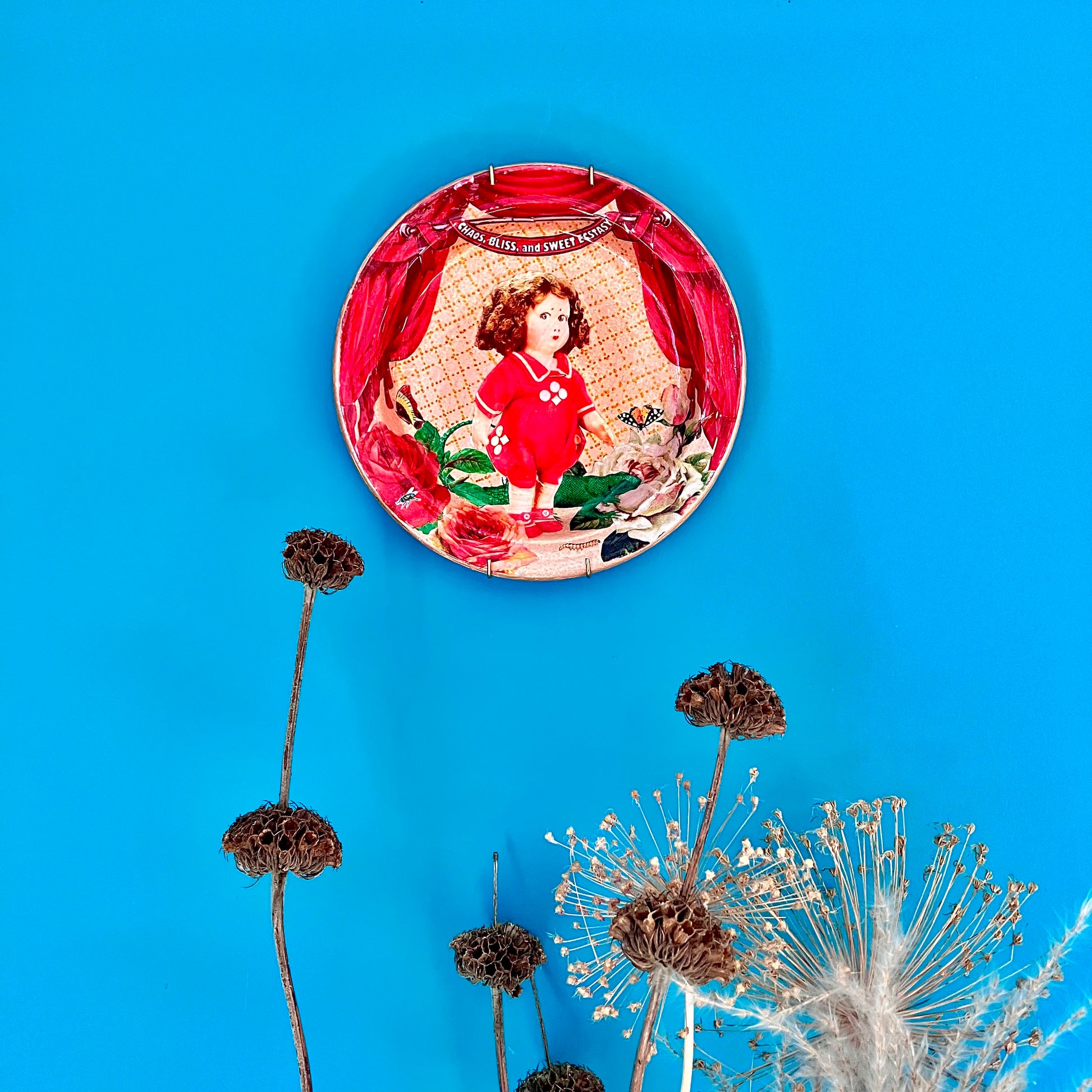 Chaos, Bliss, and Sweet Ecstasy Wall Plate by House of Frisson, hanging on a blue wallpaper.