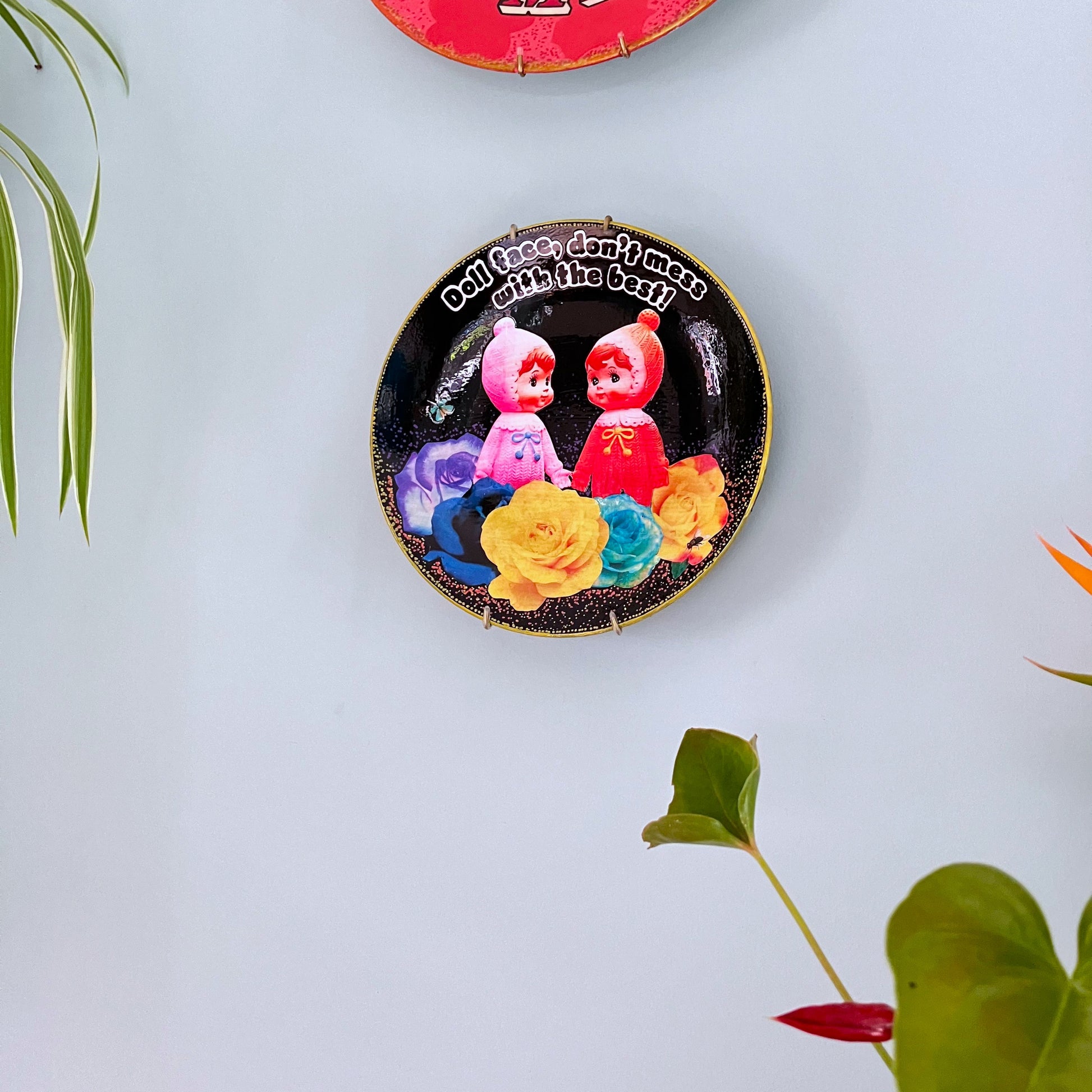 Black Upcycled Wall Plate featuring two vintage rubber dolls, surrounded by colourful roses, and a cheeky quote. Plate hanging on wall.