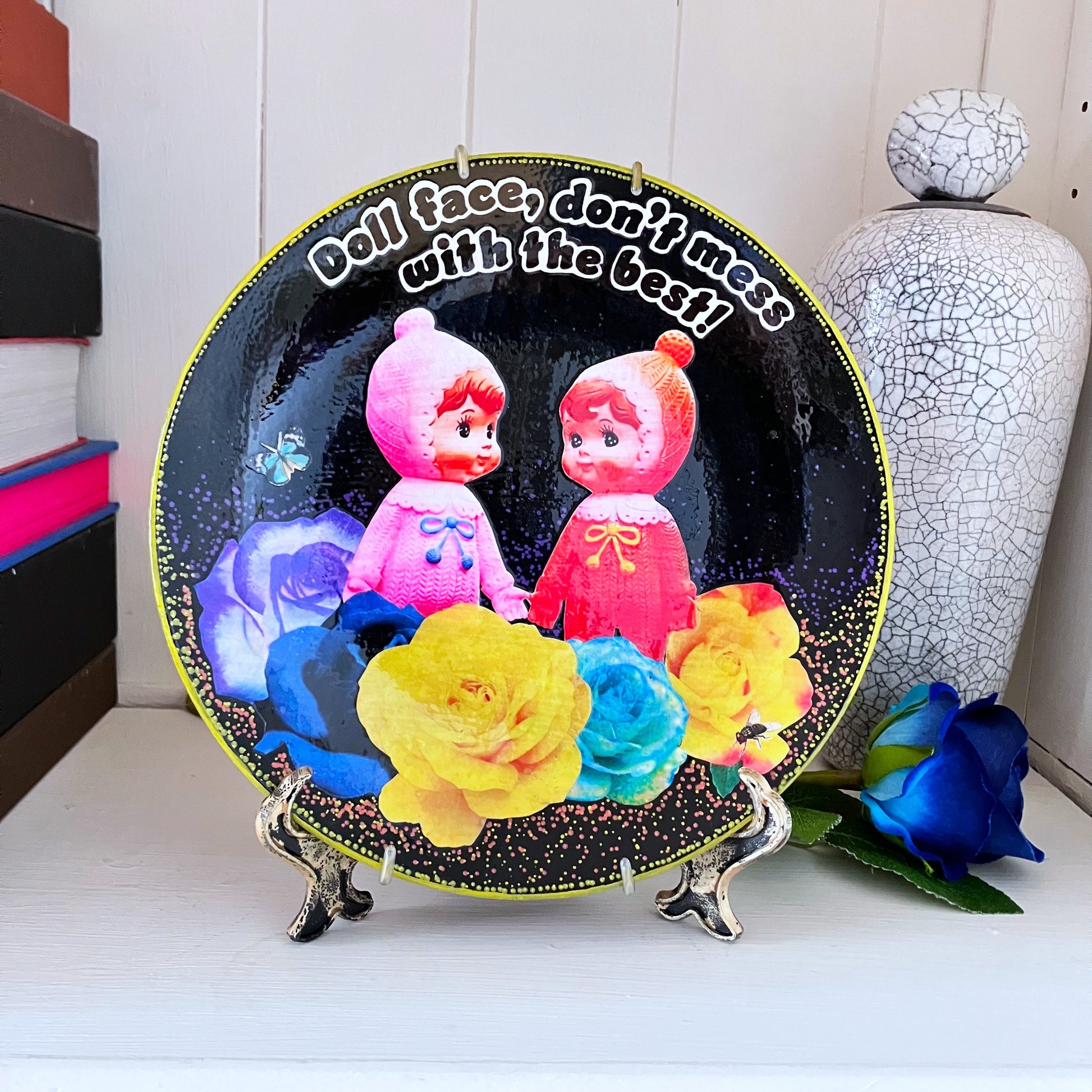 Black Upcycled Wall Plate featuring two vintage rubber dolls, surrounded by colourful roses, and a cheeky quote. Plate on a plate stand resting on a shelf.