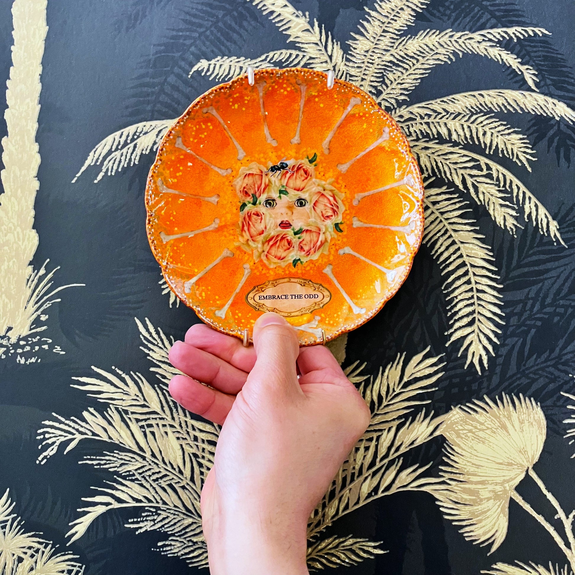 "Embrace the Odd" Orange Wall Plate by House of Frisson, featuring a one-of-a-kind paper collage of a doll's face surrounded by roses, a fly, bones, and a message box reading "Embrace the Odd". Showing someone holding the plate.