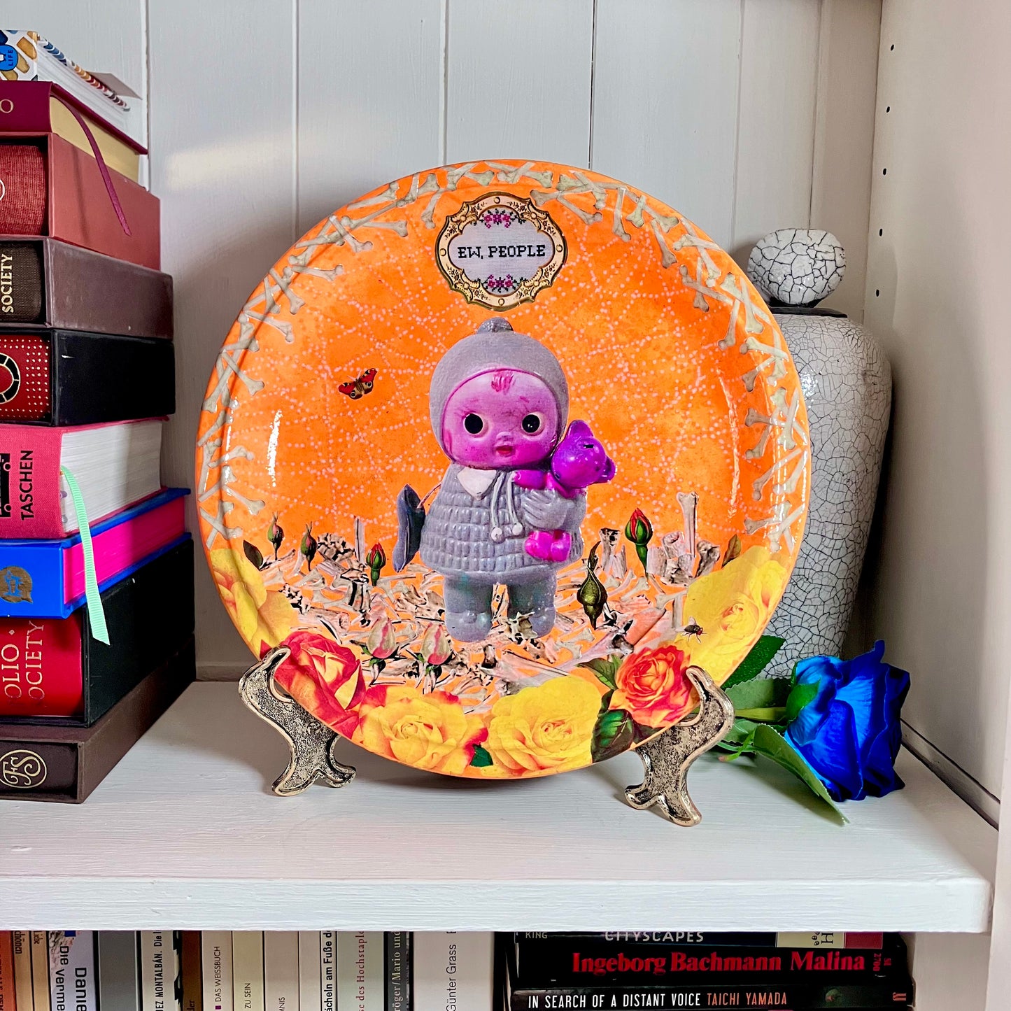 Neon orange wall plate by House of Frisson, featuring a vintage doll surrounded by bones and flowers, this plate exclaims "Ew, People" to poke fun at those who find dolls creepy. Plate on a plate stand, resting on a shelf.