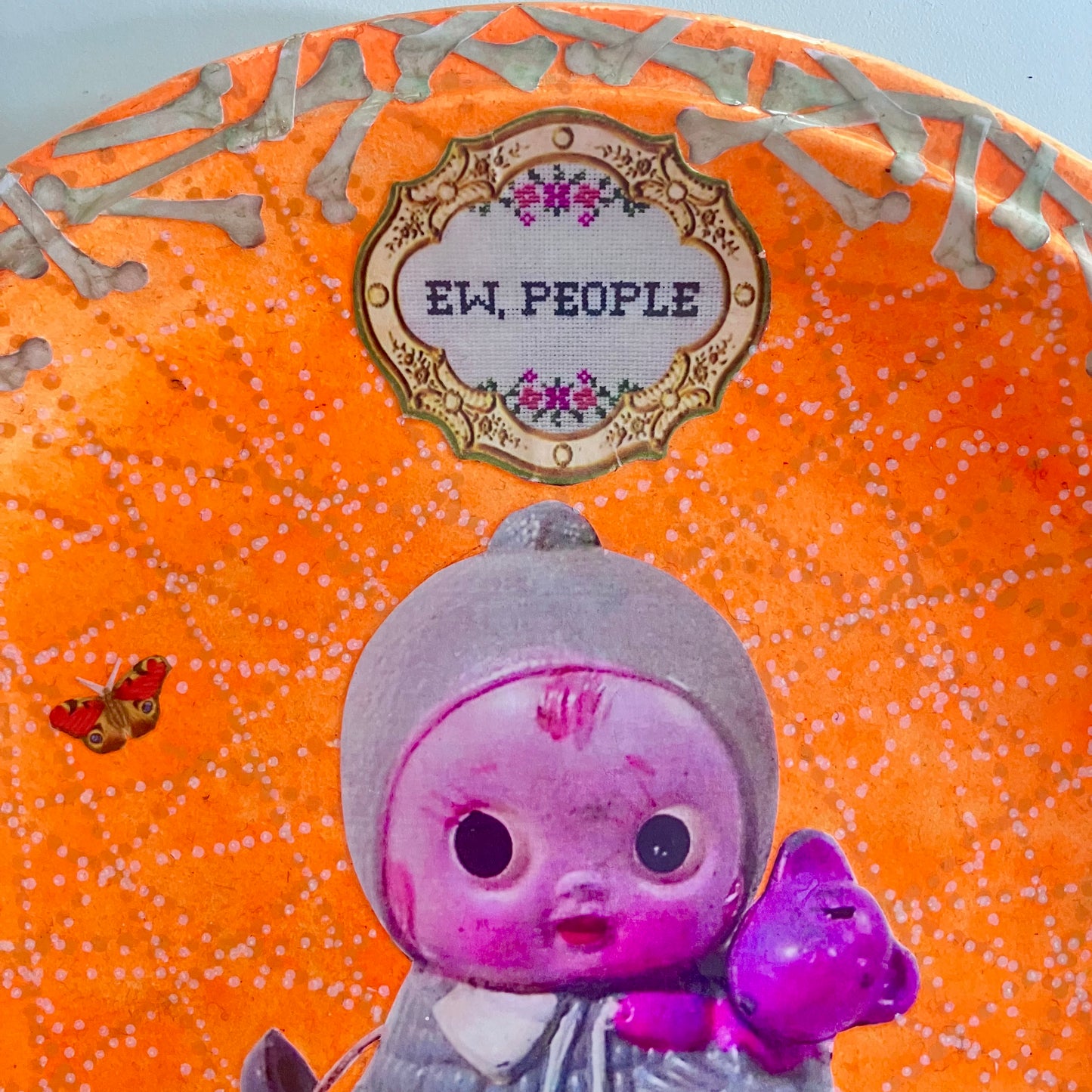 Neon orange wall plate by House of Frisson, featuring a vintage doll surrounded by bones and flowers, this plate exclaims "Ew, People" to poke fun at those who find dolls creepy. Details.