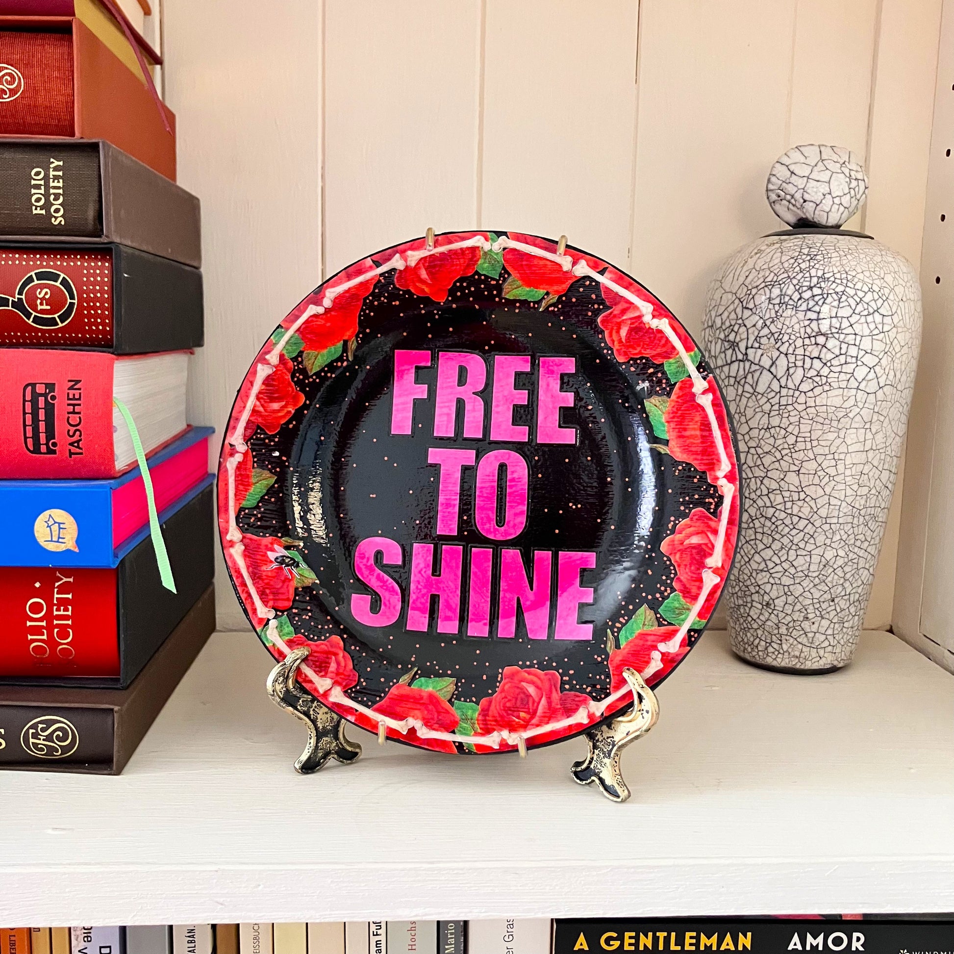 "Free To Shine" Upcycled Wall Plate by House of Frisson, featuring a unique collage of red roses and bones on a black background. Wall plate on a stand resting on a shelf.