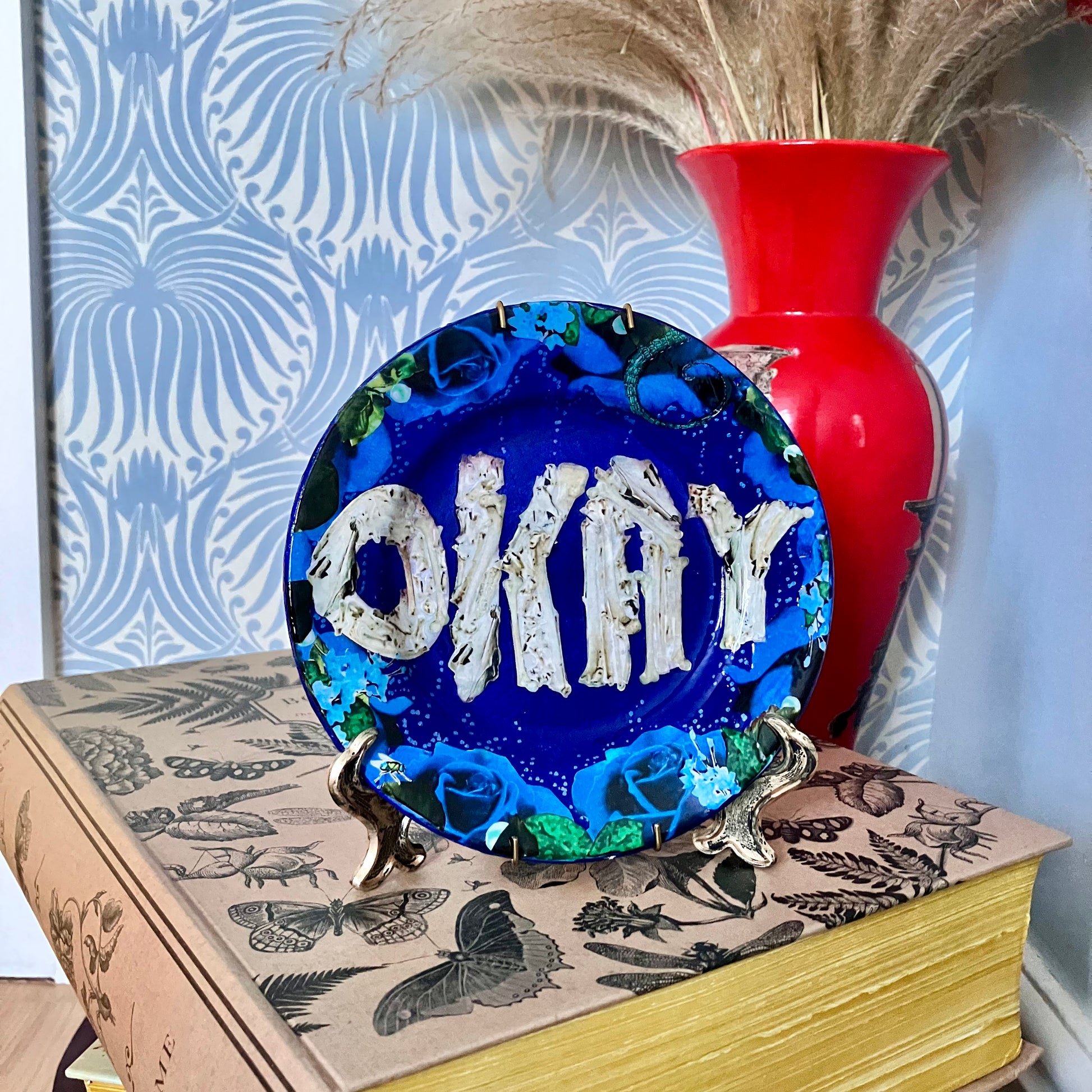 House of Frisson's "Okay" Wall Plate one-of-a-kind collage artwork on up-cycled plate. Plate on a plate stand resting on a book.