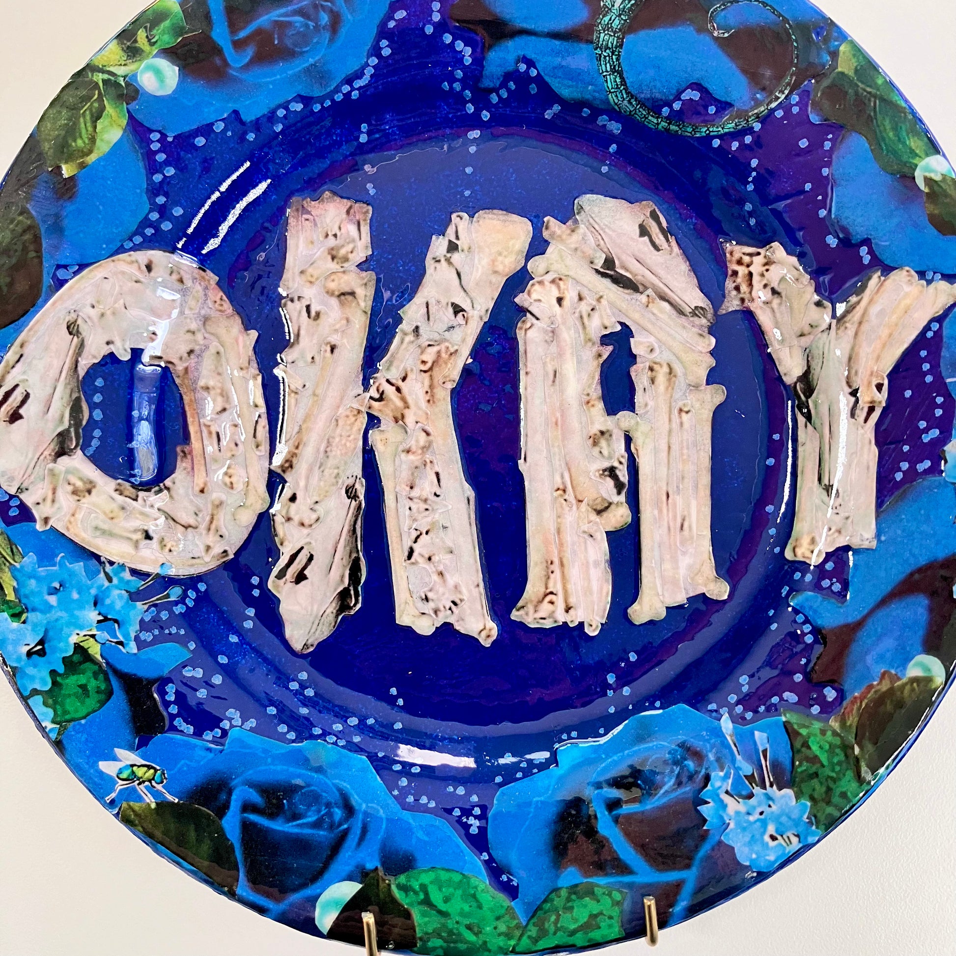 House of Frisson's "Okay" Wall Plate one-of-a-kind collage artwork on up-cycled plate. Closeup detail of the collage showing the word "Okay" written with bones, framed by blue roses, on a blue background.