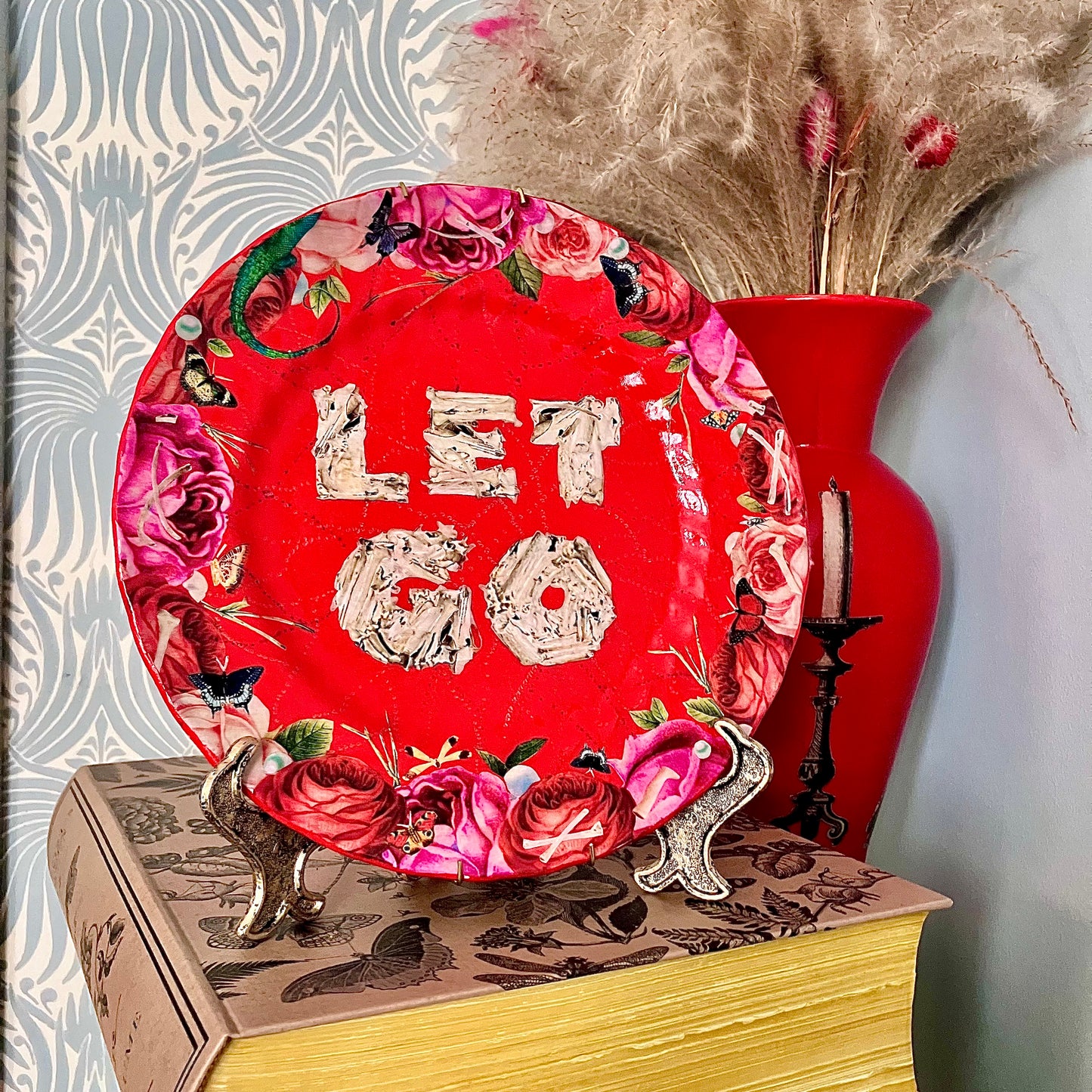 "Let Go" Wall Plate by House of Frisson on a plate stand resting on a book.