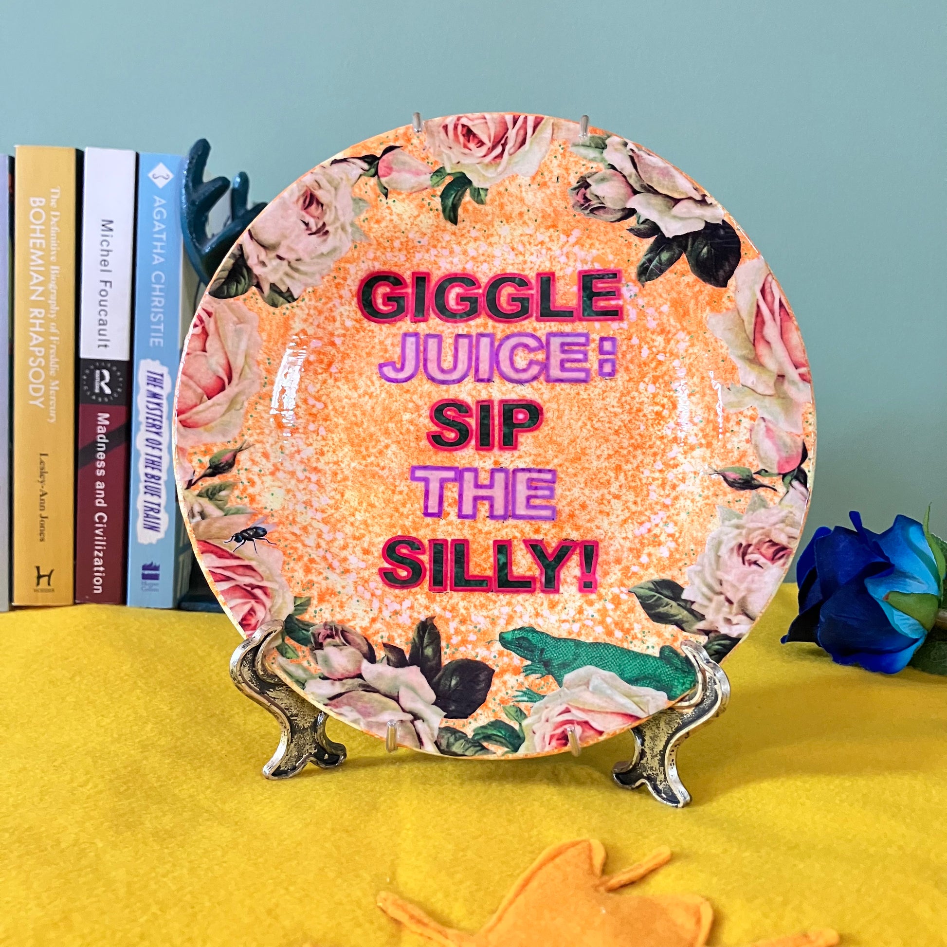 "Giggle Juice: Sip The Silly!" Orange Upcycled Wall Plate by House of Frisson, featuring the phrase "Giggle Juice: Sip The Silly!" and some roses and a lizard. Showing plate on a plate stand, resting on a table.