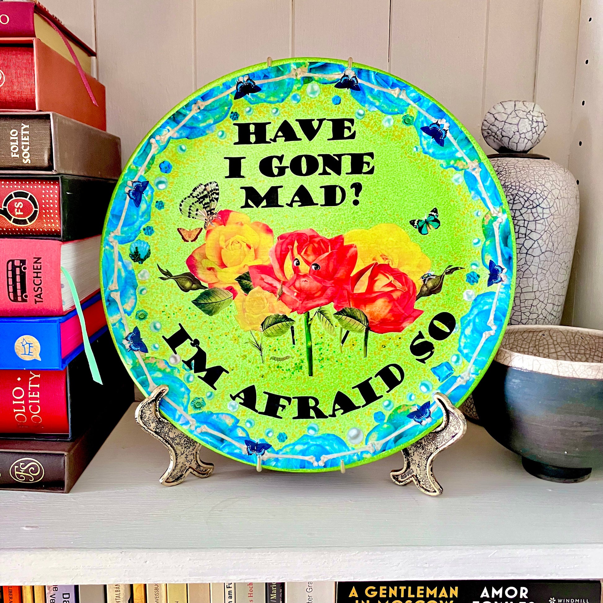 "Have I Gone Mad? I'm Afraid So" Wall Plate by House of Frisson on a plate stand resting on a bookshelf.