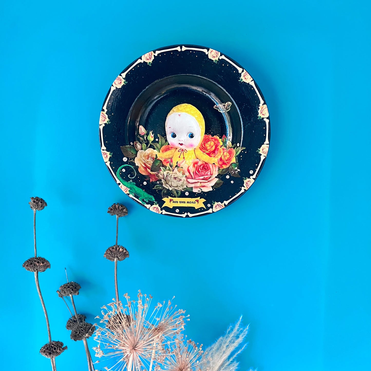 Black upcycled wall plate from House of Frisson, featuring a yellow celluloid doll surrounded by roses and pearls, in a frame of bones. Plate hanging on a blue wall.