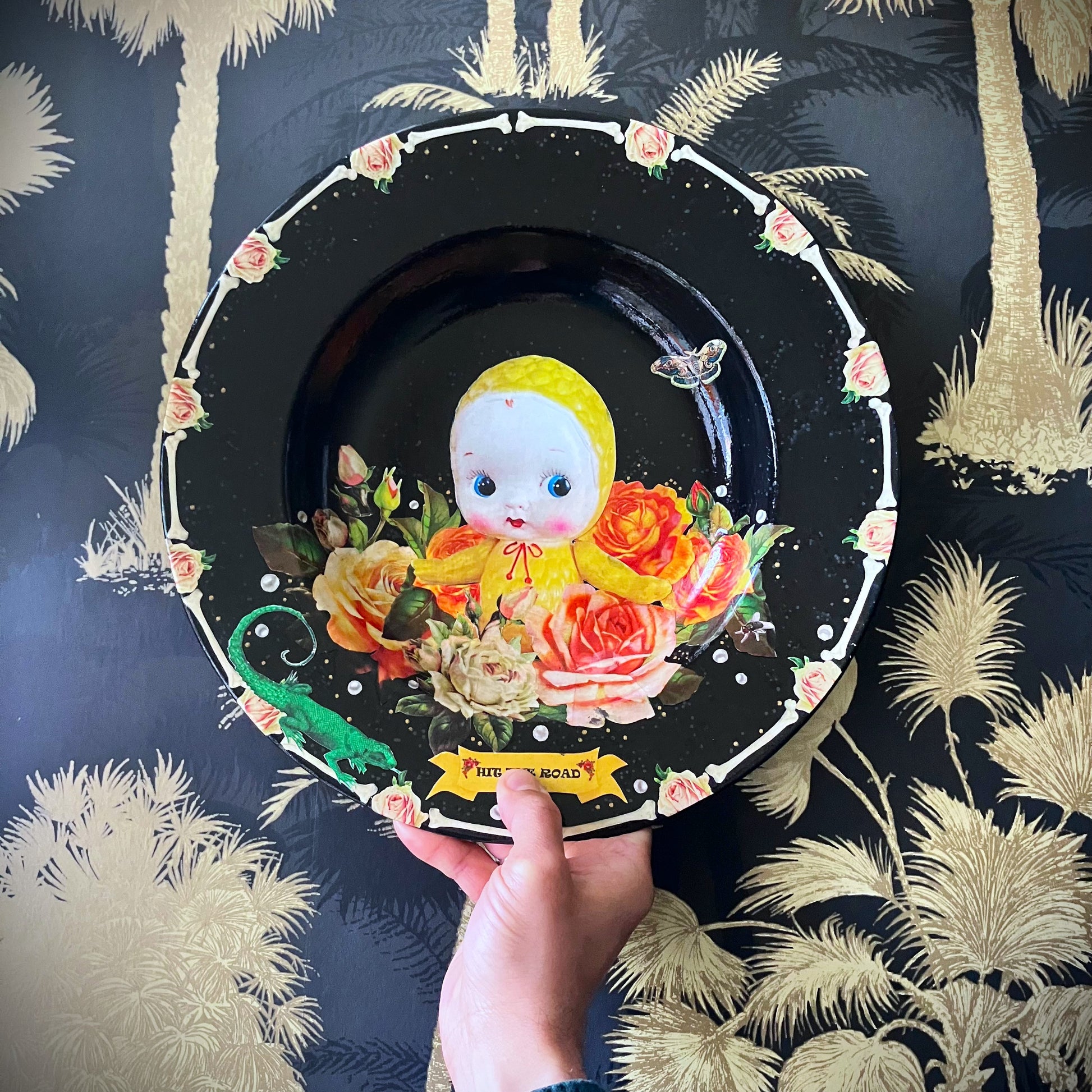 Black upcycled wall plate from House of Frisson, featuring a yellow celluloid doll surrounded by roses and pearls, in a frame of bones.