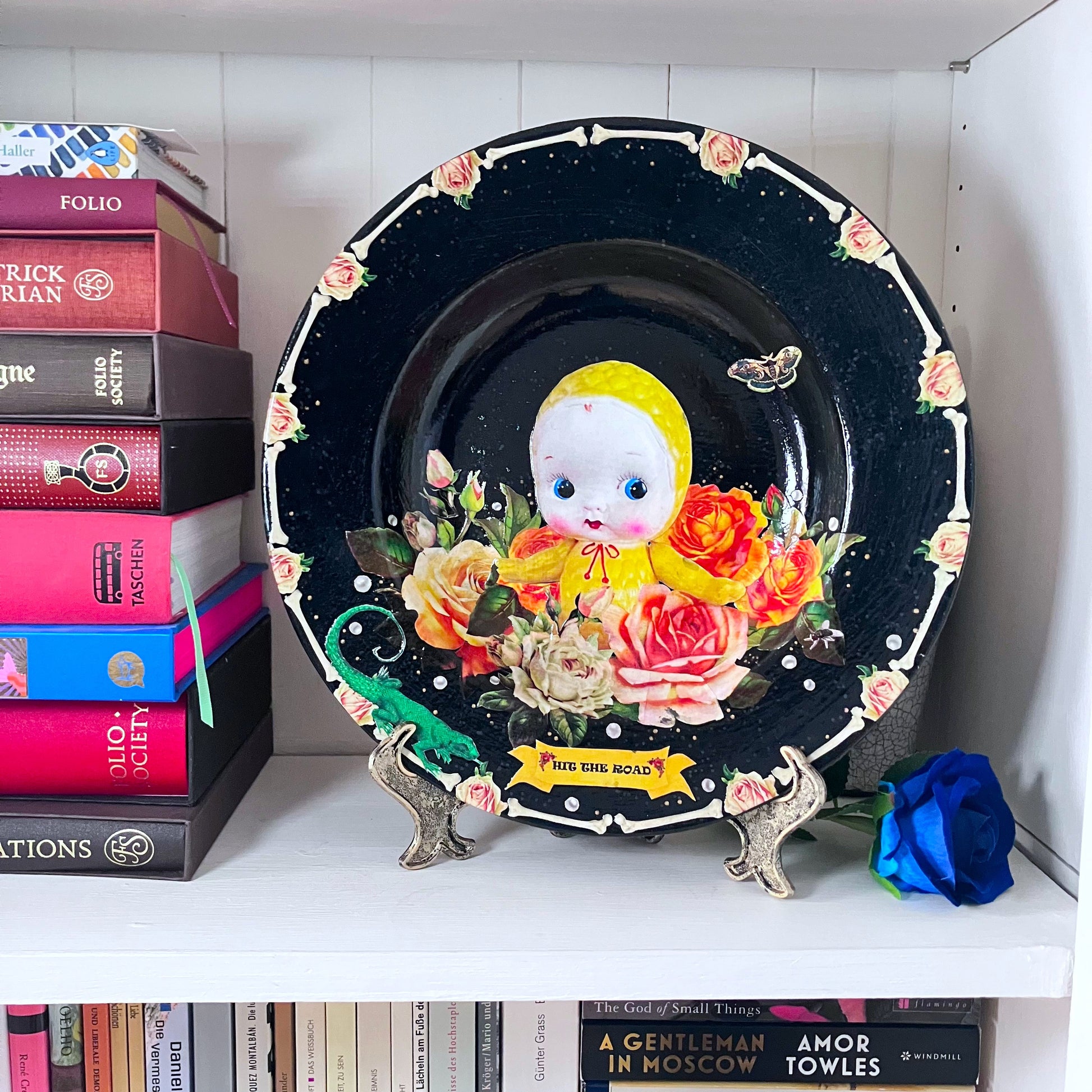 Black upcycled wall plate from House of Frisson, featuring a yellow celluloid doll surrounded by roses and pearls, in a frame of bones. late on a plate stand resting on a shelf.