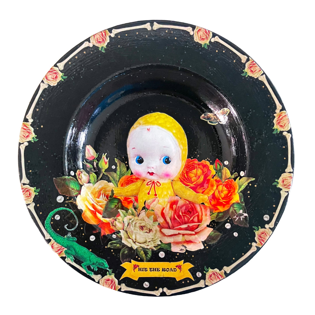 Black upcycled wall plate from House of Frisson, featuring a yellow celluloid doll surrounded by roses and pearls, in a frame of bones.