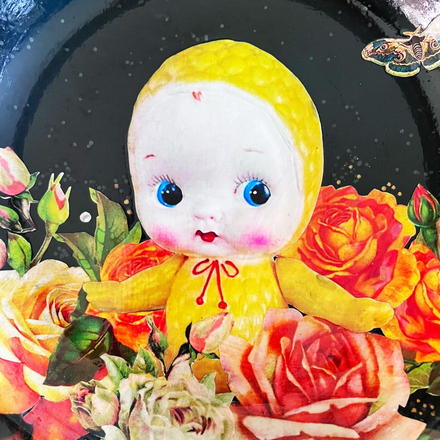 Black upcycled wall plate from House of Frisson, featuring a yellow celluloid doll surrounded by roses and pearls, in a frame of bones. Details.