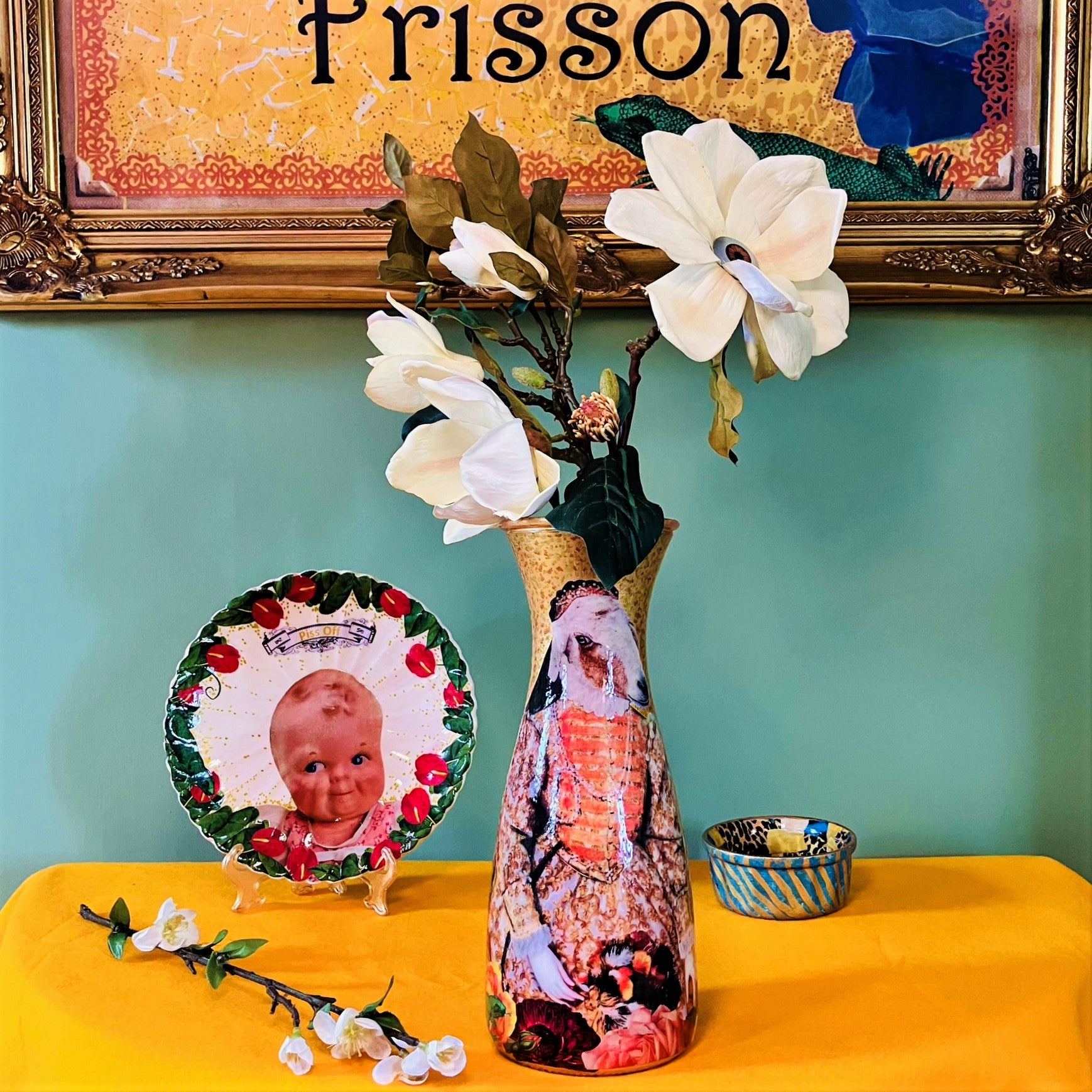 "Be Nice Or Get Lost" Flower Vase by House of Frisson, featuring collage of Queen Elizabeth I with a goat head, on a gold background. Vase displayed on a table with other objects.