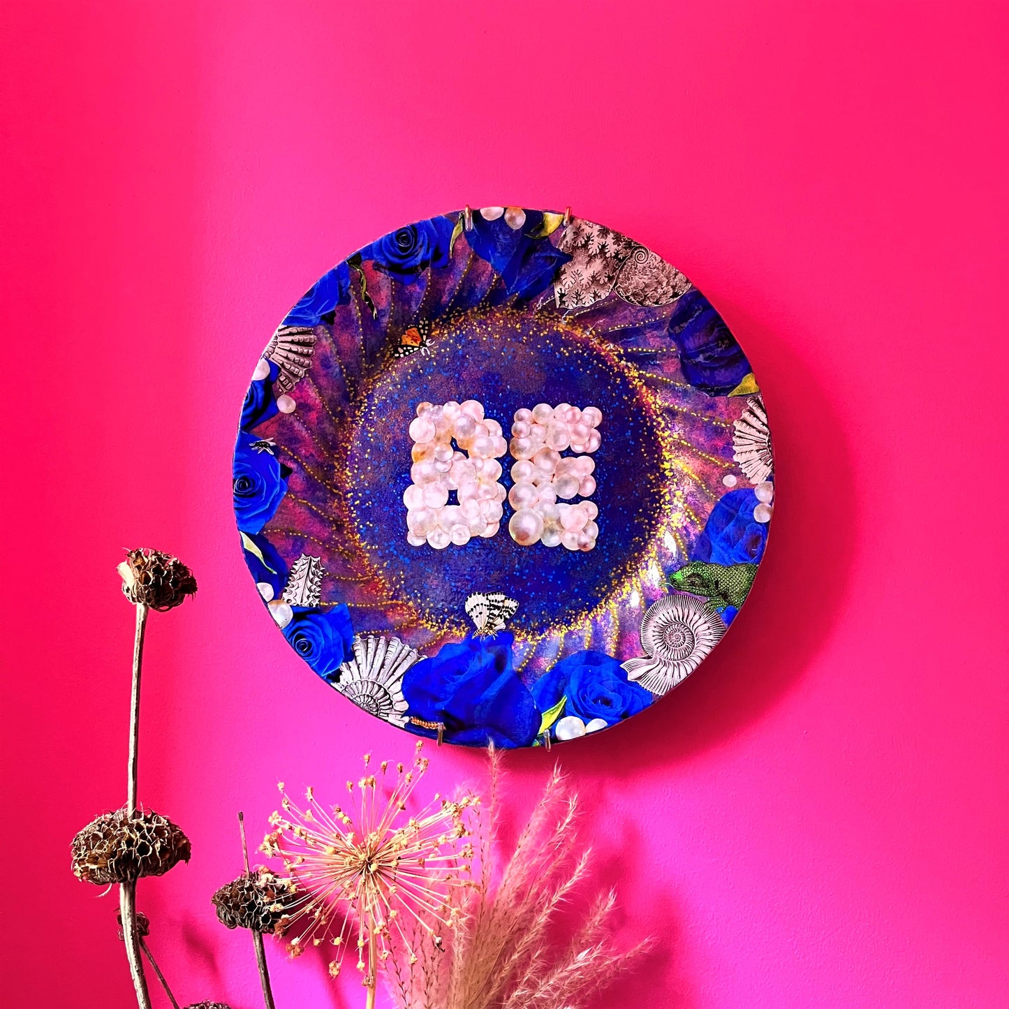 "Be" Wall Plate by House of Frisson. Featuring a collage of the word "Be" written with pearls, framed by blue roses, pearls, shells, and moths, on a blue background with golden details. Lifestyle photo of plate on a pink wall.