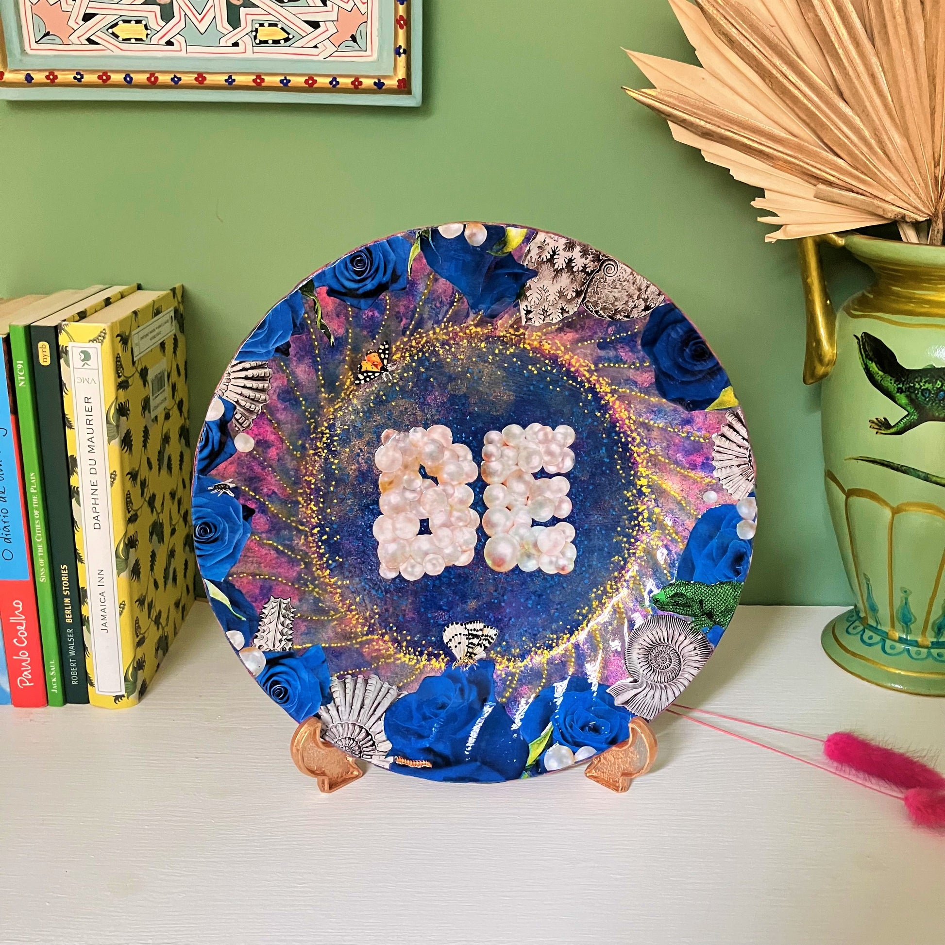 "Be" Wall Plate by House of Frisson. Featuring a collage of the word "Be" written with pearls, framed by blue roses, pearls, shells, and moths, on a blue background with golden details. Plate on a plate stand, resting on a shelf.
