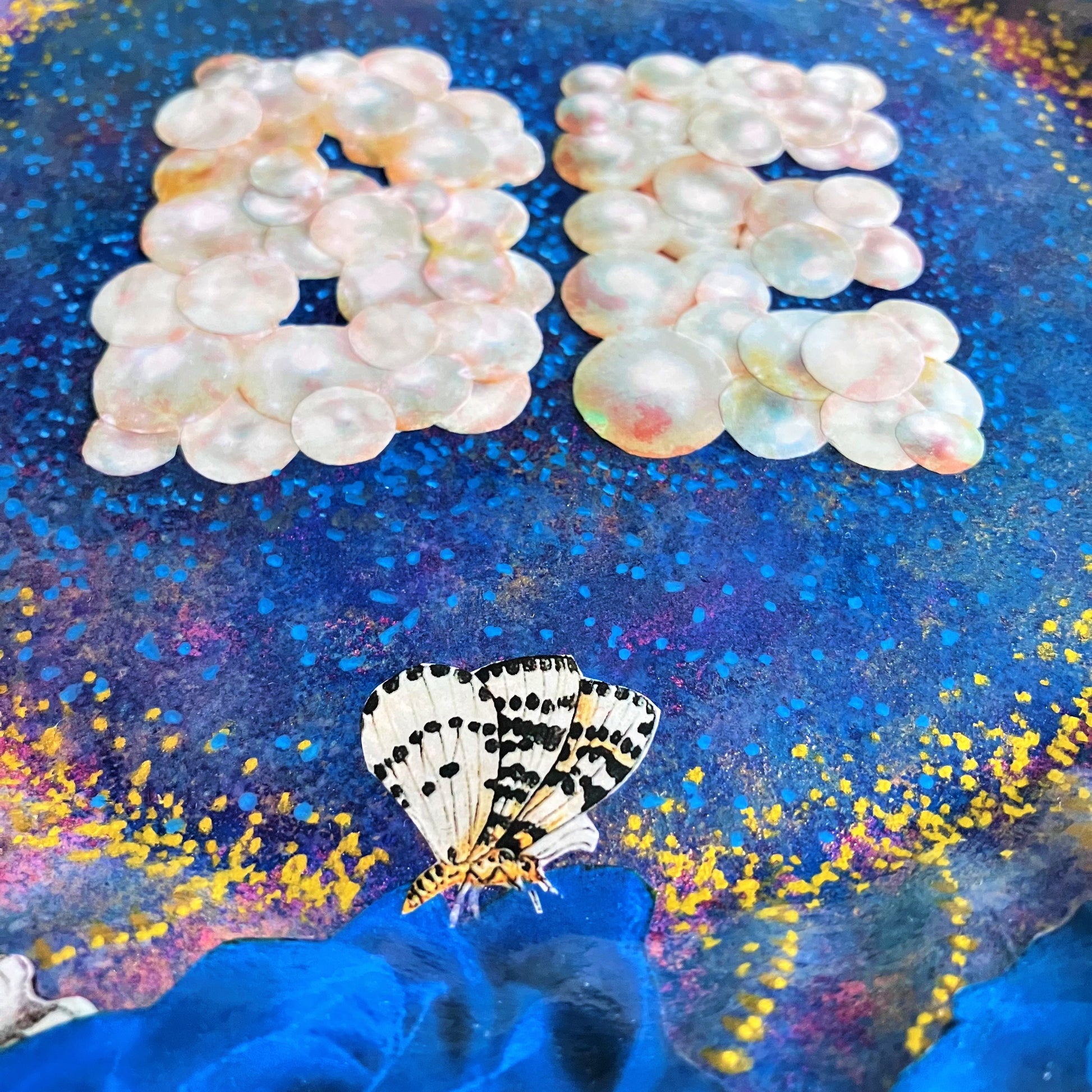 "Be" Wall Plate by House of Frisson. Featuring a collage of the word "Be" written with pearls, framed by blue roses, pearls, shells, and moths, on a blue background with golden details. Closeup detail showing the blue roses and moths.