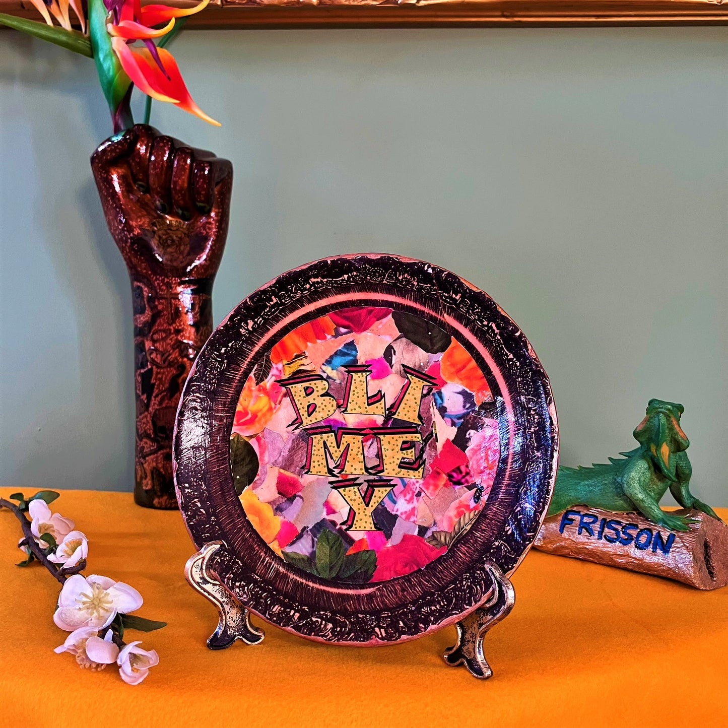 "Blimey" Wall Plate by House of Frisson, featuring a collage of word "blimey" framed with a vintage looking frame, with colourful background. Plate on a plate stand, resting on a table.