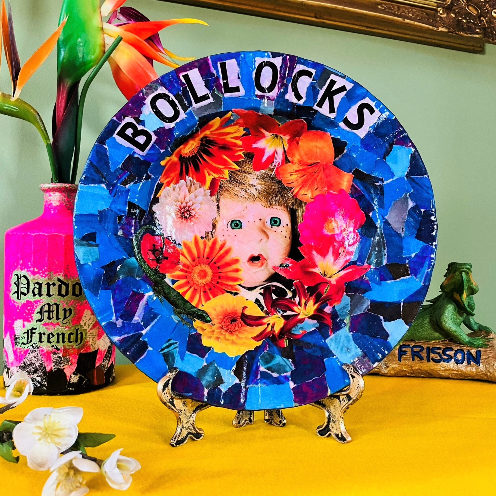 "Bollocks" Wall Plate by House of Frisson, featuring a collage of a vintage male doll surrounded by flowers and a lizard, on a blue background. Plate on a plate stand, resting on a table.