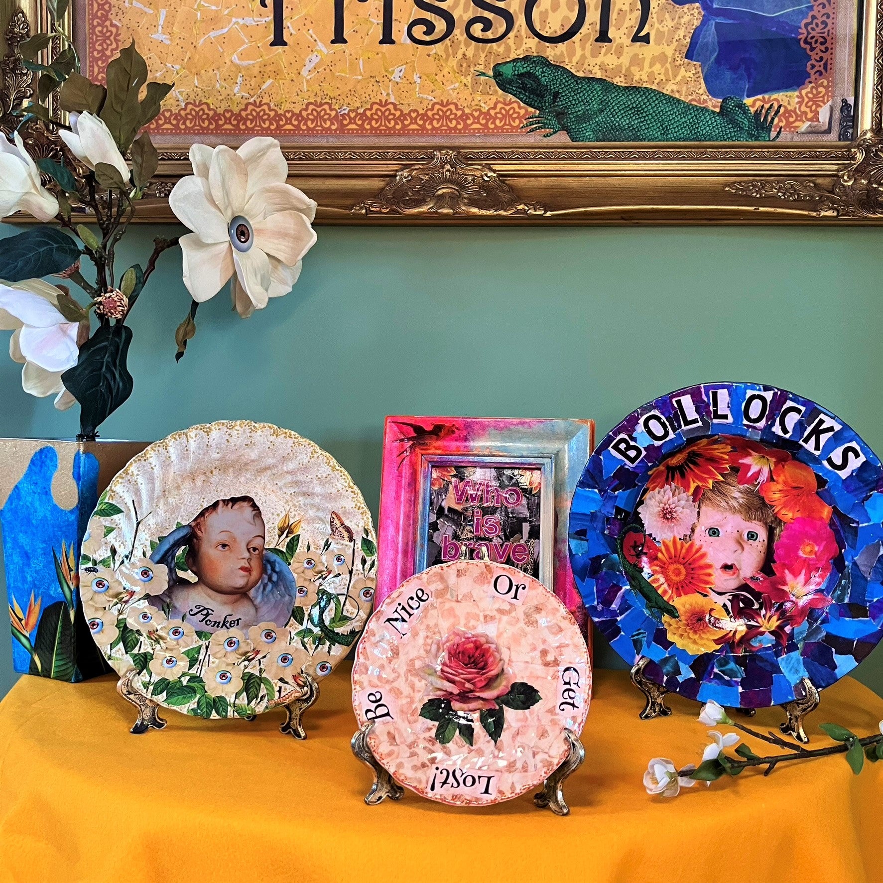 "Bollocks" Wall Plate by House of Frisson, featuring a collage of a vintage male doll surrounded by flowers and a lizard, on a blue background. Plate on plate stand, resting on a table with other plates.
