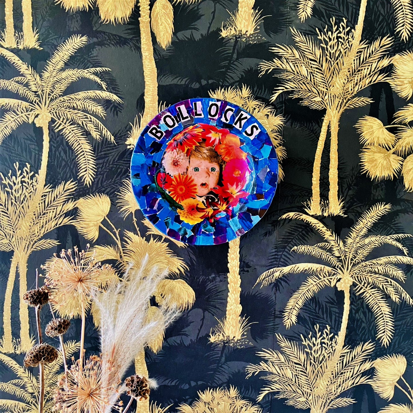 "Bollocks" Wall Plate by House of Frisson, featuring a collage of a vintage male doll surrounded by flowers and a lizard, on a blue background. Plate hanging on a wall papered wall.