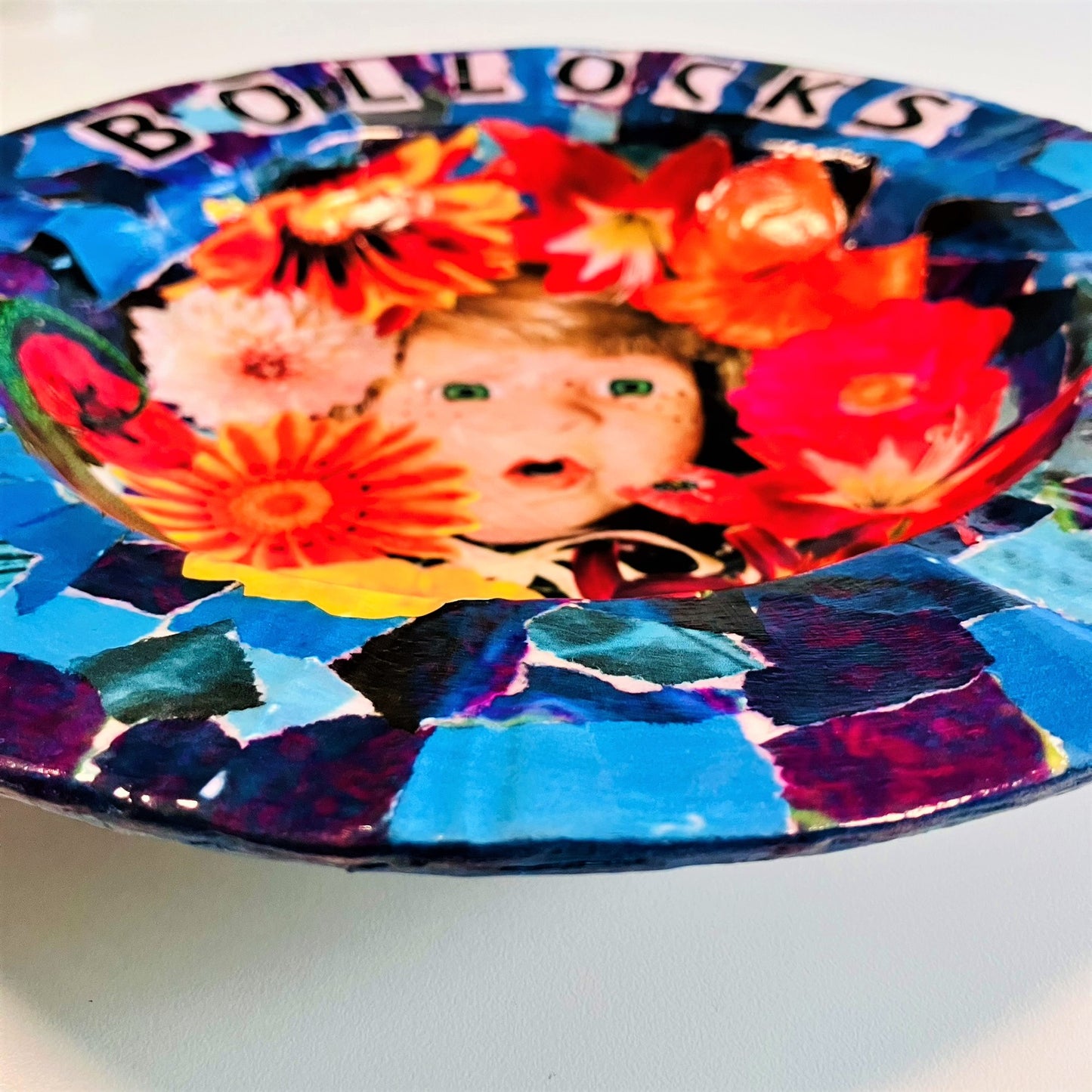 "Bollocks" Wall Plate by House of Frisson, featuring a collage of a vintage male doll surrounded by flowers and a lizard, on a blue background. Closeup details showing blue background.