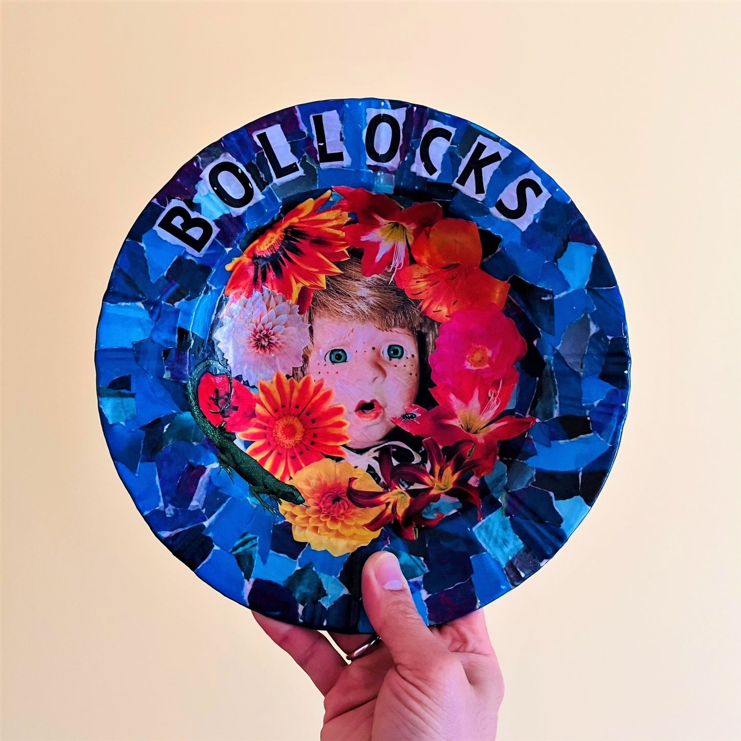 "Bollocks" Wall Plate by House of Frisson, featuring a collage of a vintage male doll surrounded by flowers and a lizard, on a blue background. Holding plate.