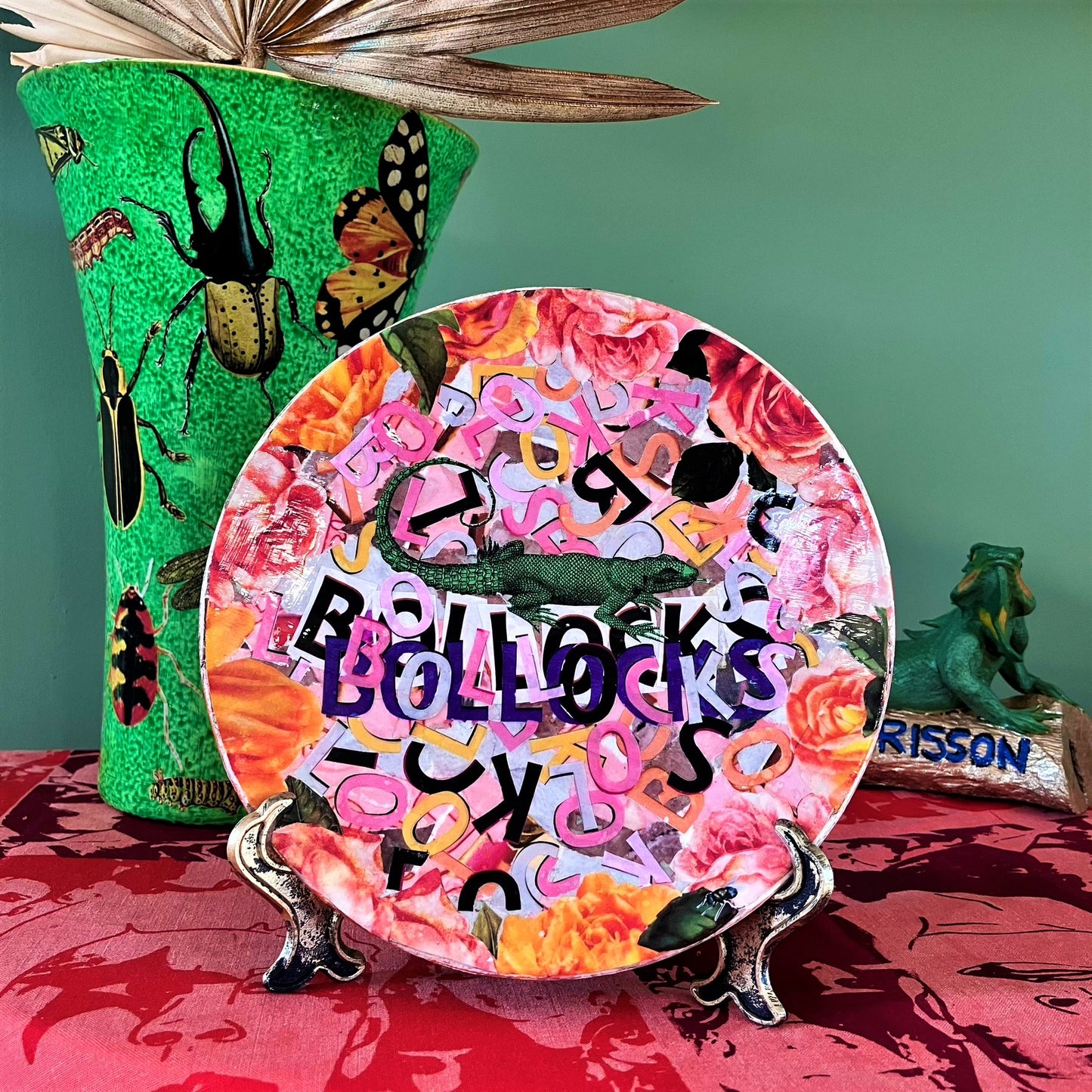 "Bollocks" Wall Plate by House of Frisson, featuring a collage of colourful letters forming the word "bollocks", and a lizard, framed by roses. Plate on a plate stand, resting on a table.