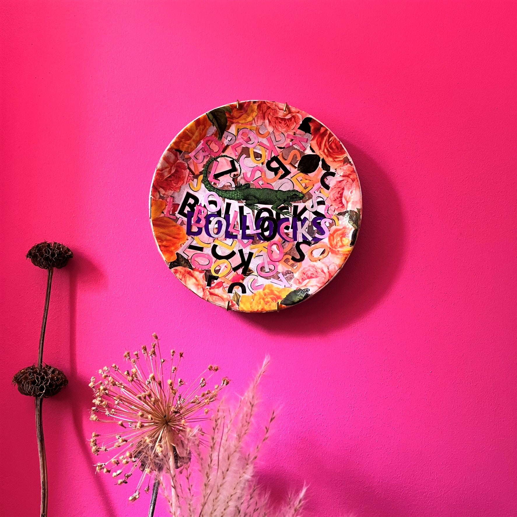 "Bollocks" Wall Plate by House of Frisson, featuring a collage of colourful letters forming the word "bollocks", and a lizard, framed by roses. Plate hanging on a pink wall.