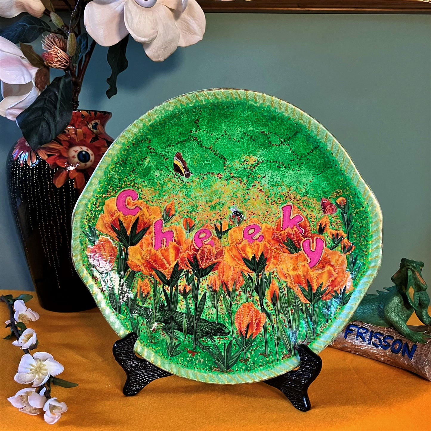 "Cheeky" Wall Plate by House of Frisson, featuring a collage with letters coming out of flowers forming the word "cheeky", and a lizard, on a green background. Showing plate on a plate stand, resting on a table.