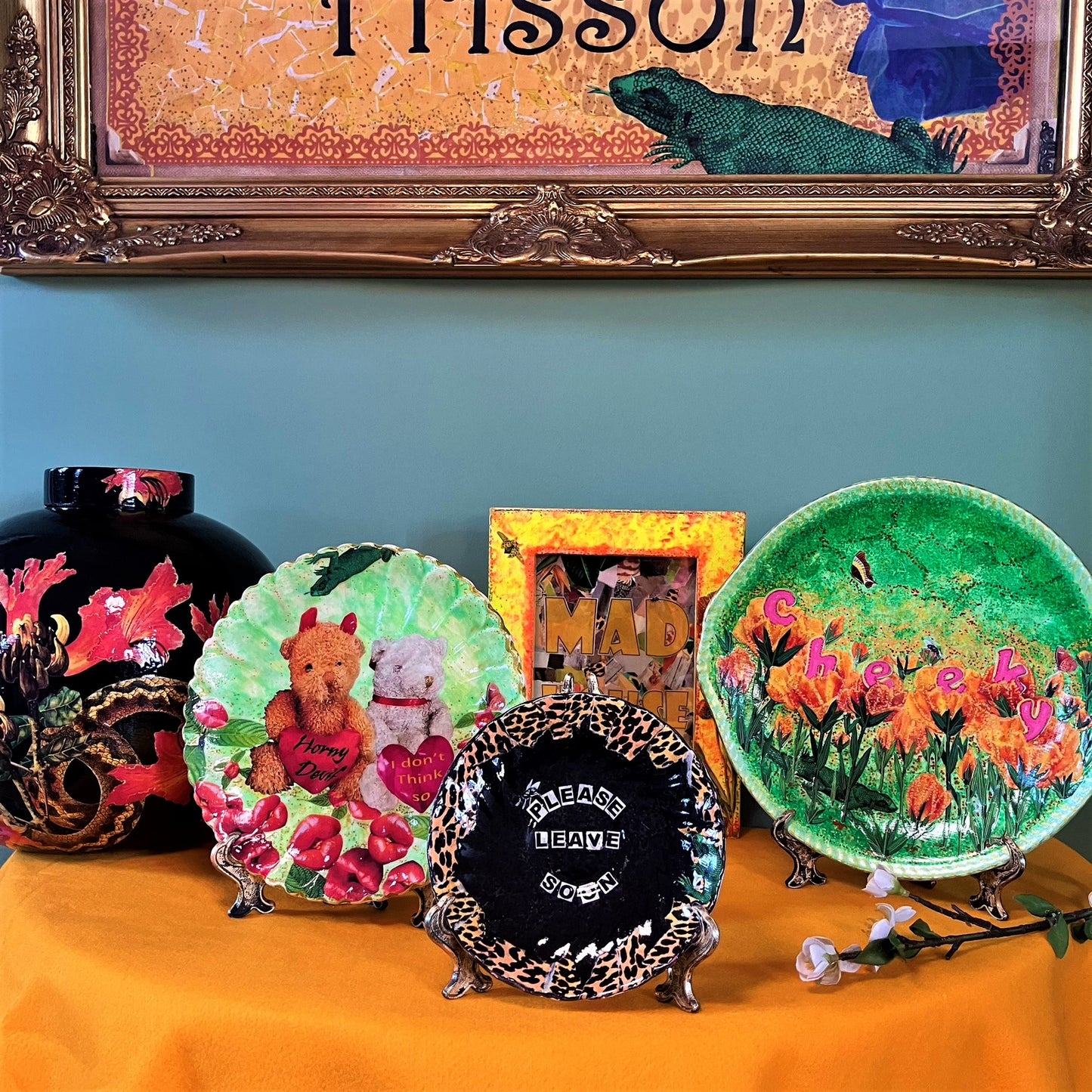 "Cheeky" Wall Plate by House of Frisson, featuring a collage with letters coming out of flowers forming the word "cheeky", and a lizard, on a green background. Plate on a plate stand resting on a table with other plates.