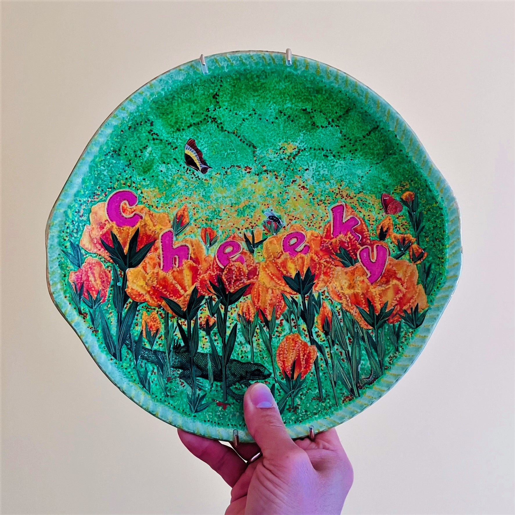 "Cheeky" Wall Plate by House of Frisson, featuring a collage with letters coming out of flowers forming the word "cheeky", and a lizard, on a green background. Holding plate.