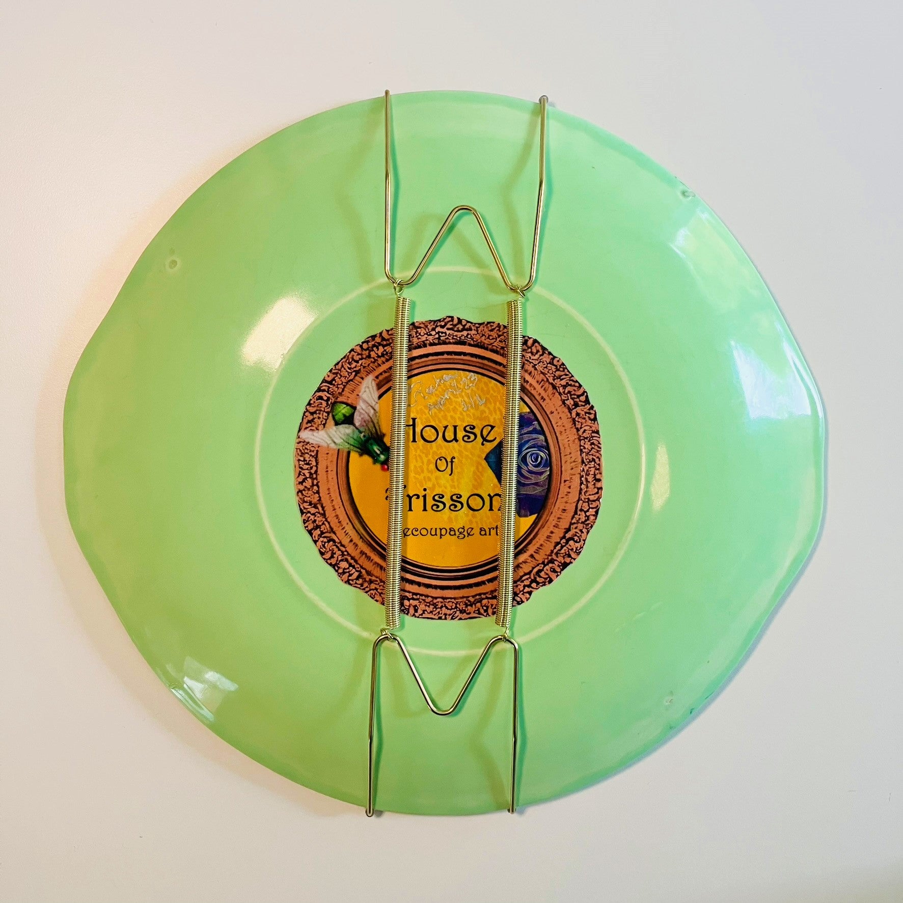"Cheeky" Wall Plate by House of Frisson, featuring a collage with letters coming out of flowers forming the word "cheeky", and a lizard, on a green background. Showing back of the plate with attached plate hanger.