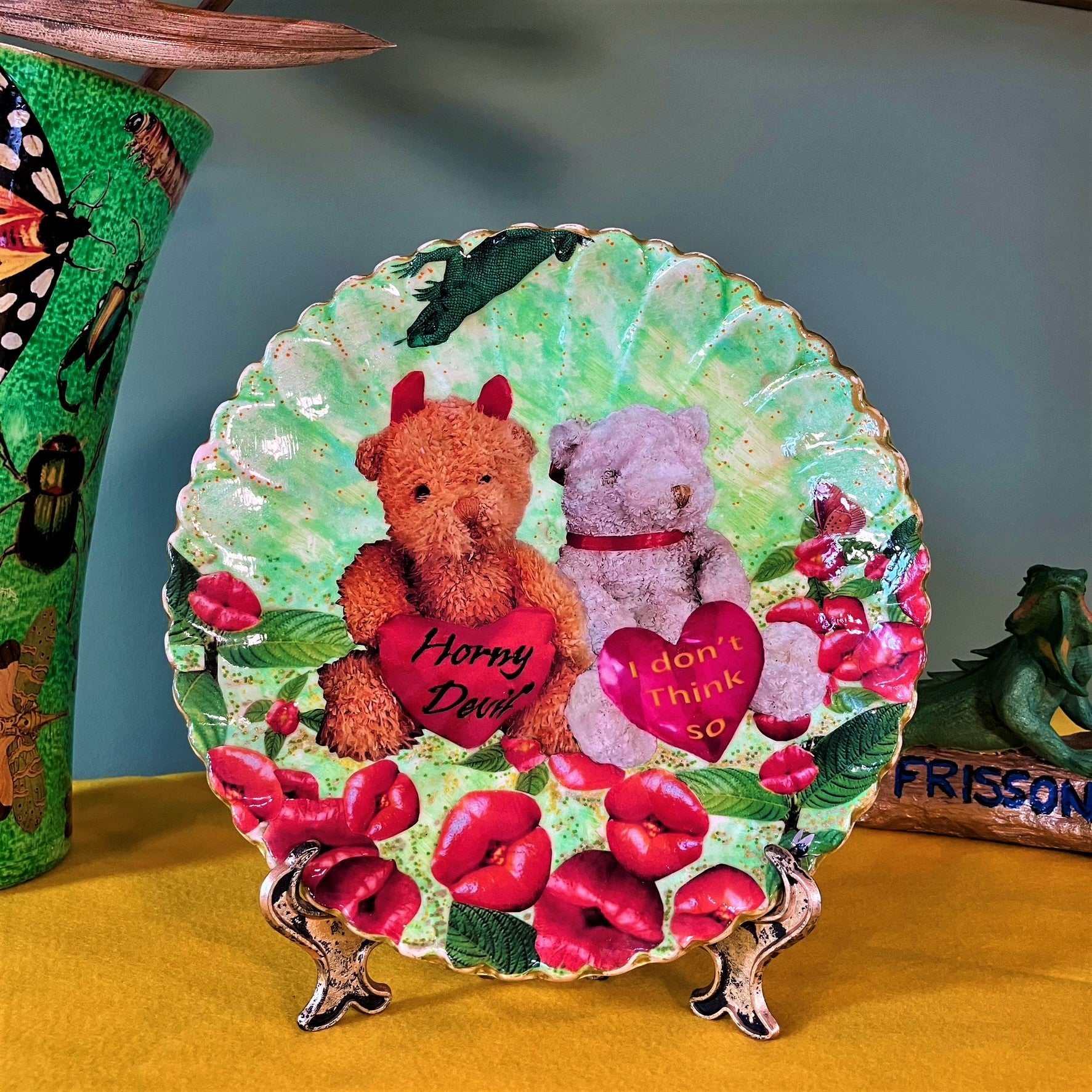 "Horny Devil. I Don't Think So" Wall Plate by House of Frisson, featuring a collage of two teddy bears holding hearts with messages, among lip-shaped flowers, on a green background. Plate on a plate stand, resting on a table.
