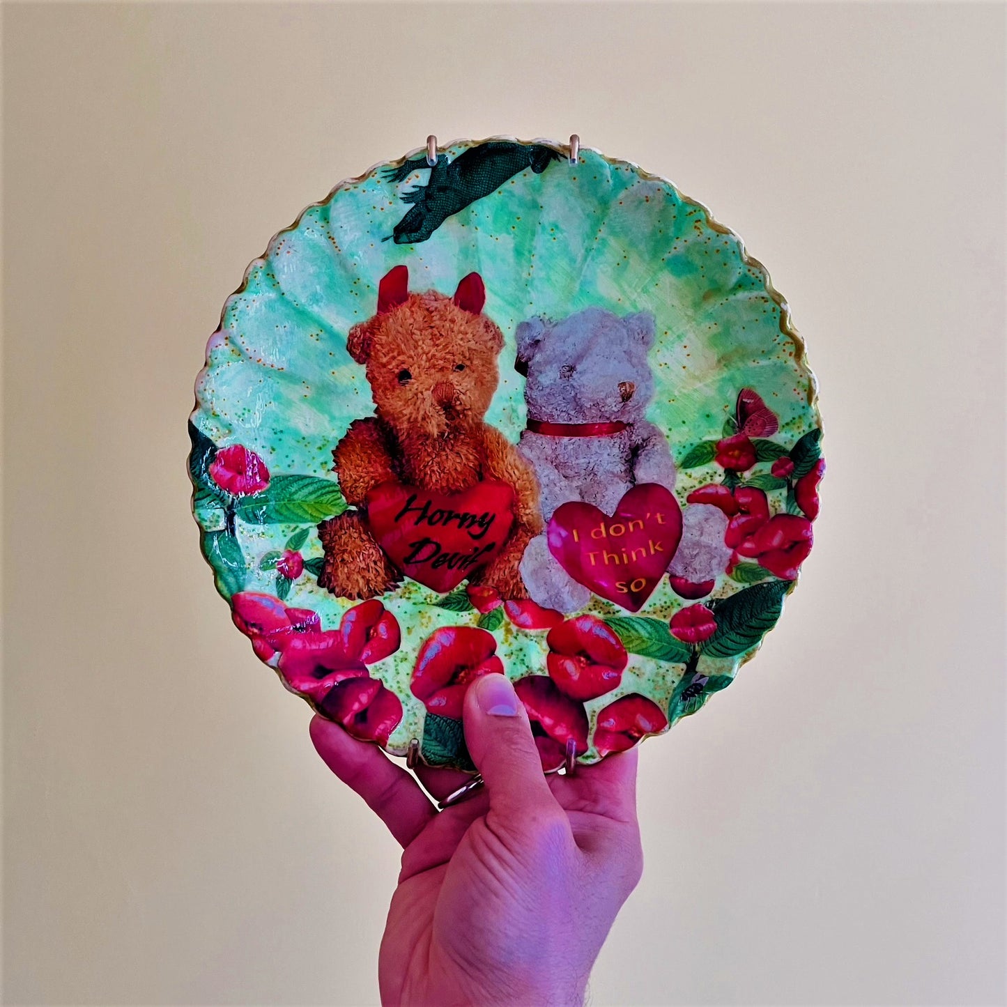 "Horny Devil. I Don't Think So" Wall Plate by House of Frisson, featuring a collage of two teddy bears holding hearts with messages, among lip-shaped flowers, on a green background. Holding the plate.