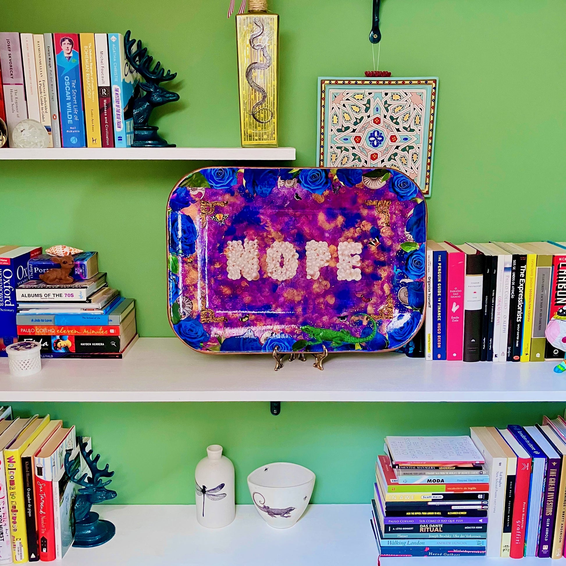 "Nope" Wall Plate by House of Frisson. Featuring the word "nope" written with pearls, framed by blue roses, shells, moths, and a lizard, on a  purple, blue and golden background. Lifestyle image showing plate on a plate stand resting on a bookshelf.
