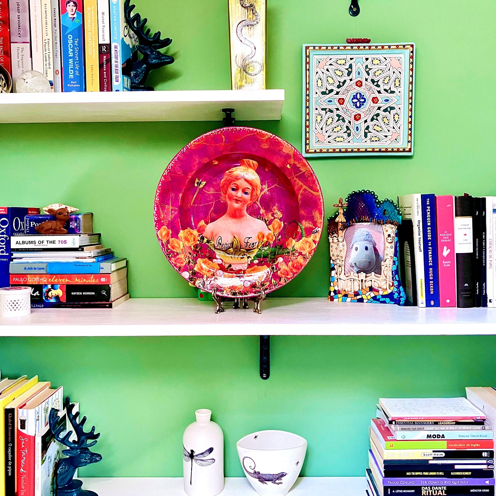 "Resist Fear" Wall Plate by House of Frisson, featuring a female bust statue with "Resist Fear" written across breasts, surrounded by flowers, and pearls, on a purple background. Showing plate on plate stand, resting on a bookshelf.