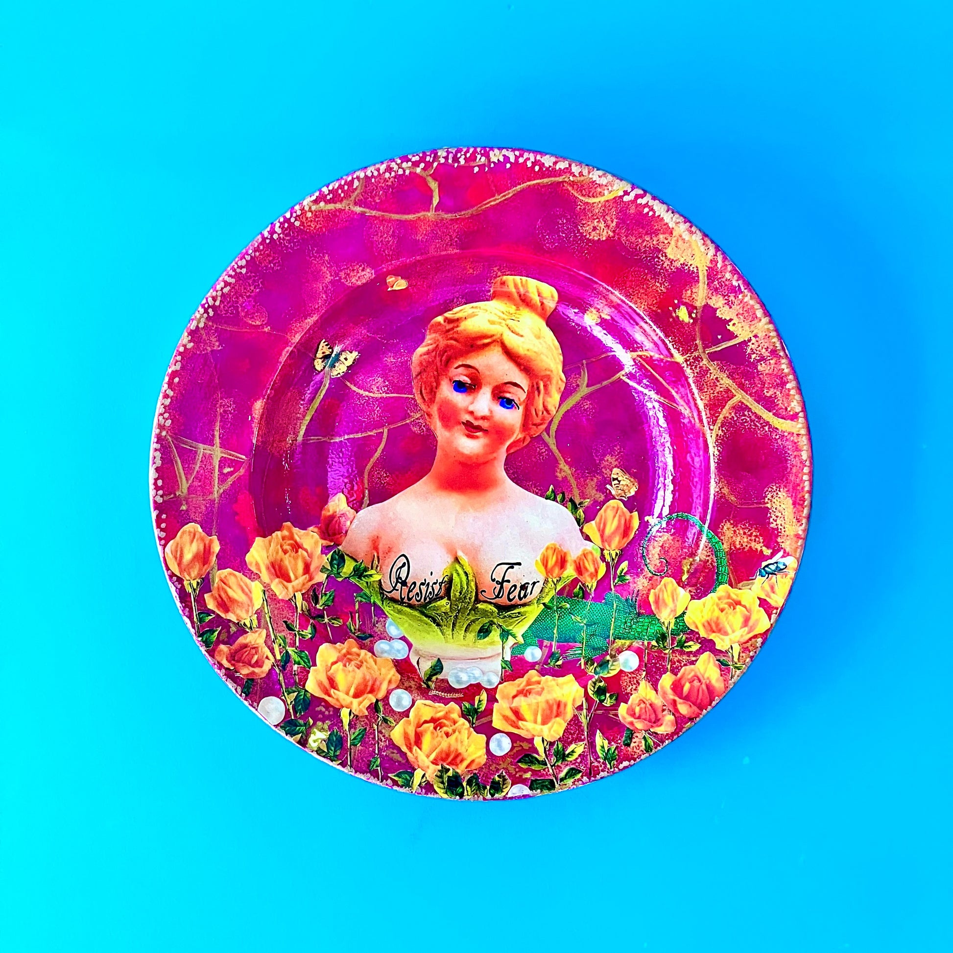 "Resist Fear" Wall Plate by House of Frisson, featuring a female bust statue with "Resist Fear" written across breasts, surrounded by flowers, and pearls, on a purple background. Showing plate on a blue wall.