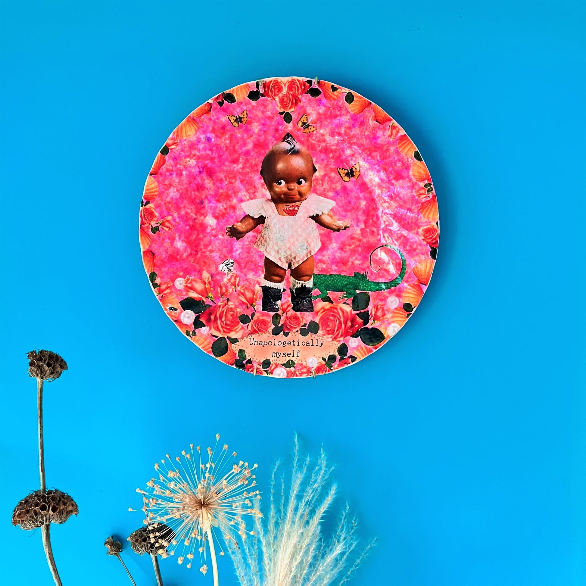 "Unapologetically Myself" Wall Plate by House of Frisson, featuring a vintage black doll surrounded by roses , pearls, moths, and a lizard, framed by shells, on a pink background. Plate on a blue wall.