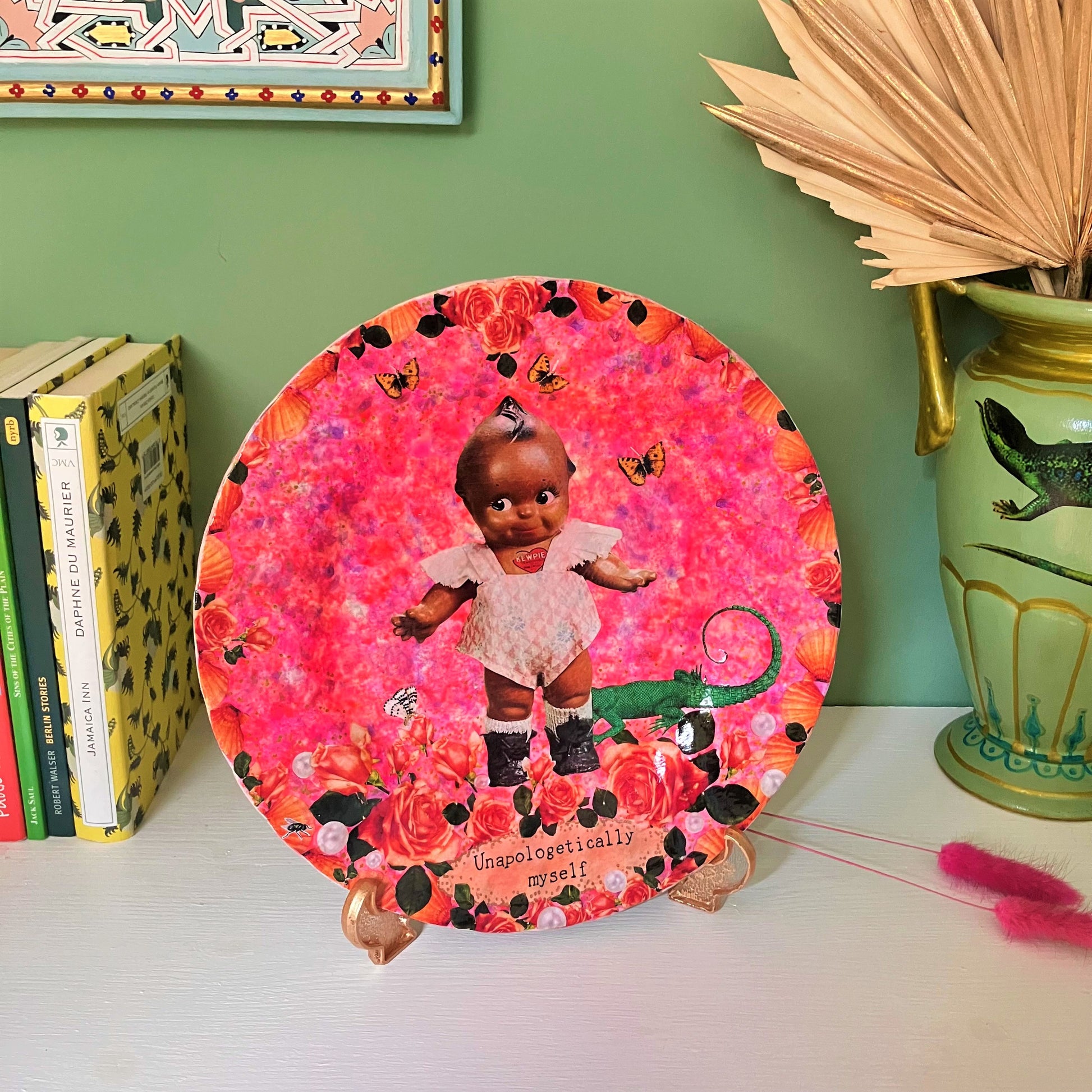 "Unapologetically Myself" Wall Plate by House of Frisson, featuring a vintage black doll surrounded by roses , pearls, moths, and a lizard, framed by shells, on a pink background. Plate on a plate stand, resting on a shelf.