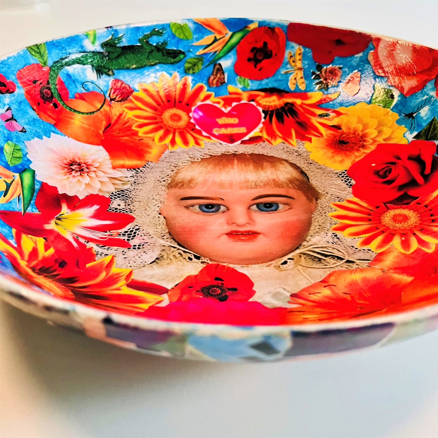 "Who Cares" Wall Plate by House of Frisson, featuring a vintage doll surrounded by flowers, moths, and a lizard, on a blue background. Showing a closeup detail of the vintage doll.