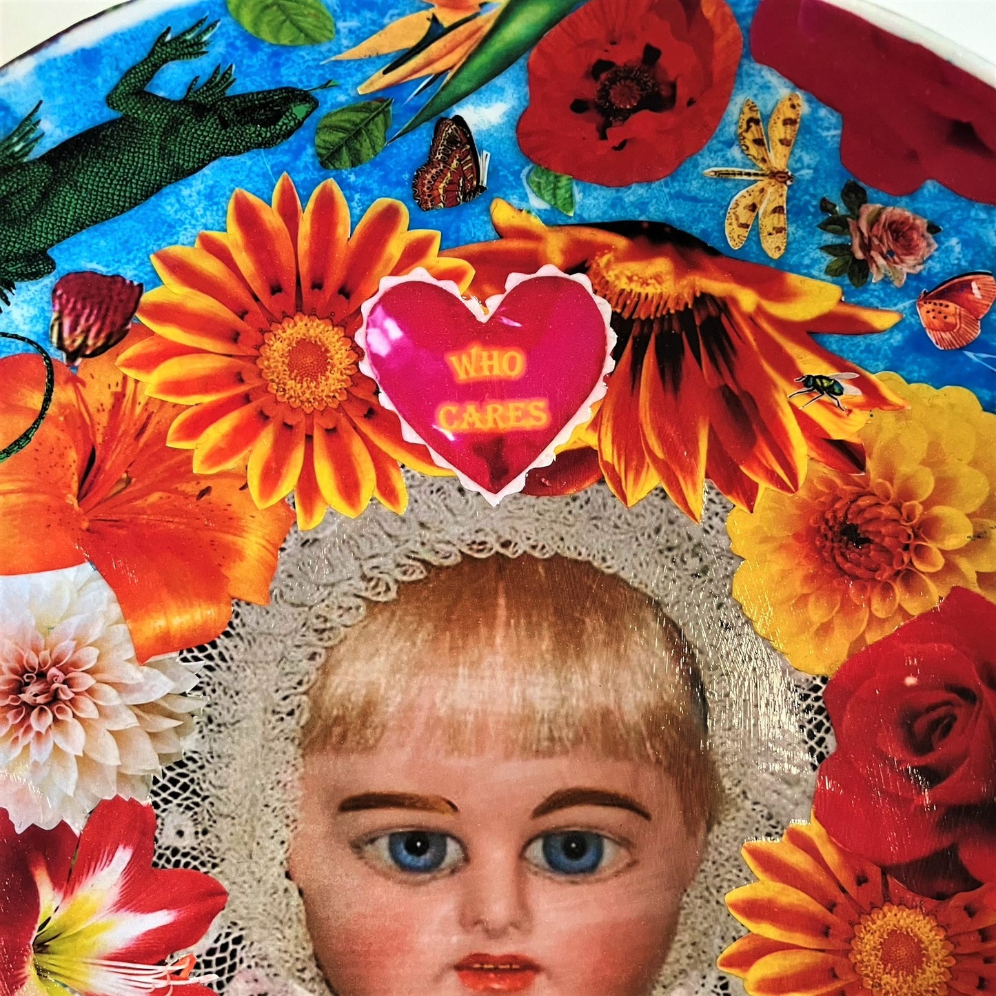 "Who Cares" Wall Plate by House of Frisson, featuring a vintage doll surrounded by flowers, moths, and a lizard, on a blue background. Showing a closeup detail of the colourful flowers and a heart with "Who Cares" written on it.